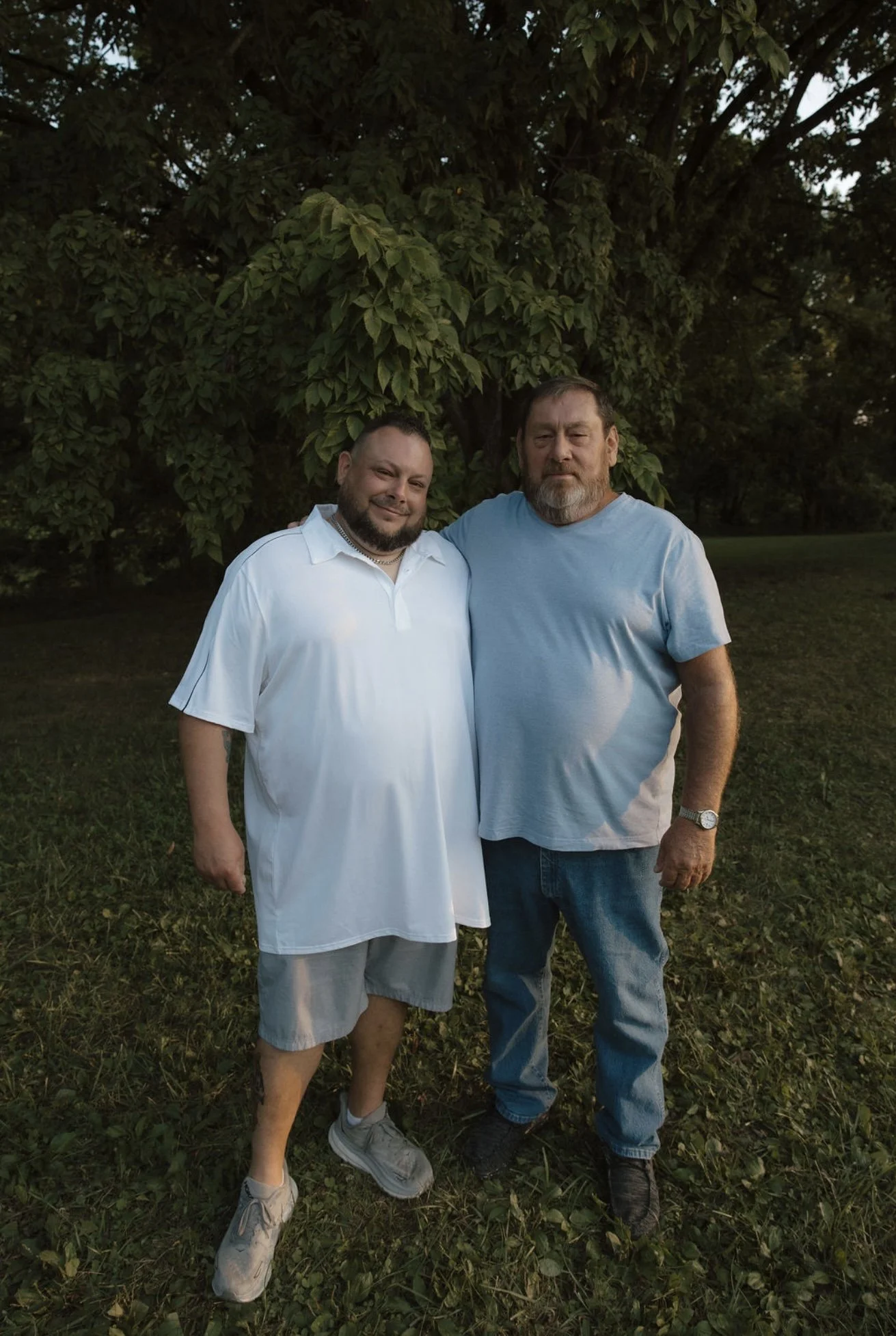 Two men standing outdoors near a tree, posing together for a photo, one smiling and wearing a white polo shirt and shorts, the other with a serious expression wearing a blue t-shirt and jeans.