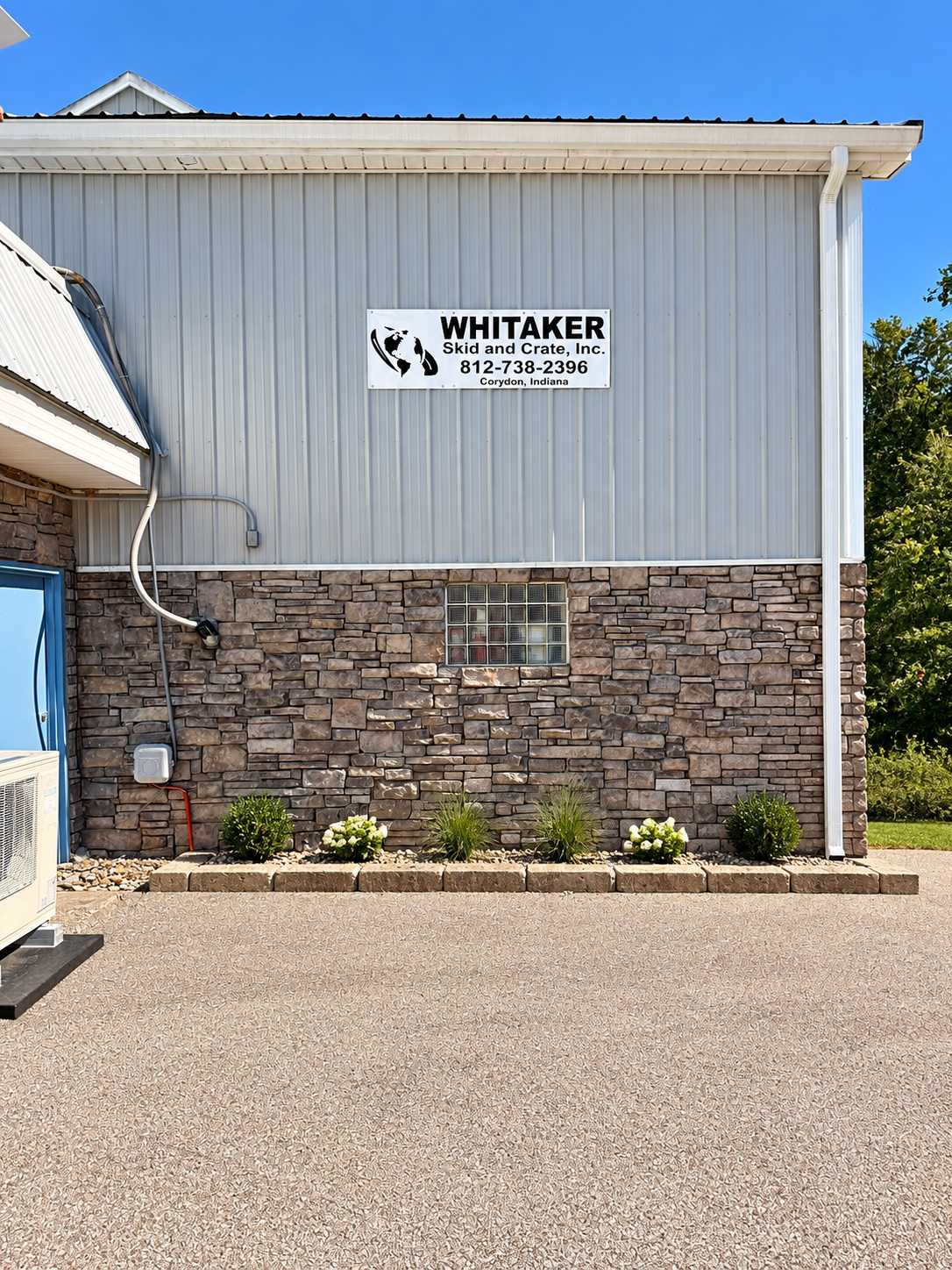 Exterior of a building with a sign for Whitaker Skid and Crate, Inc. in Corydon, Indiana, with brick and metal siding and small landscaped flower bed.