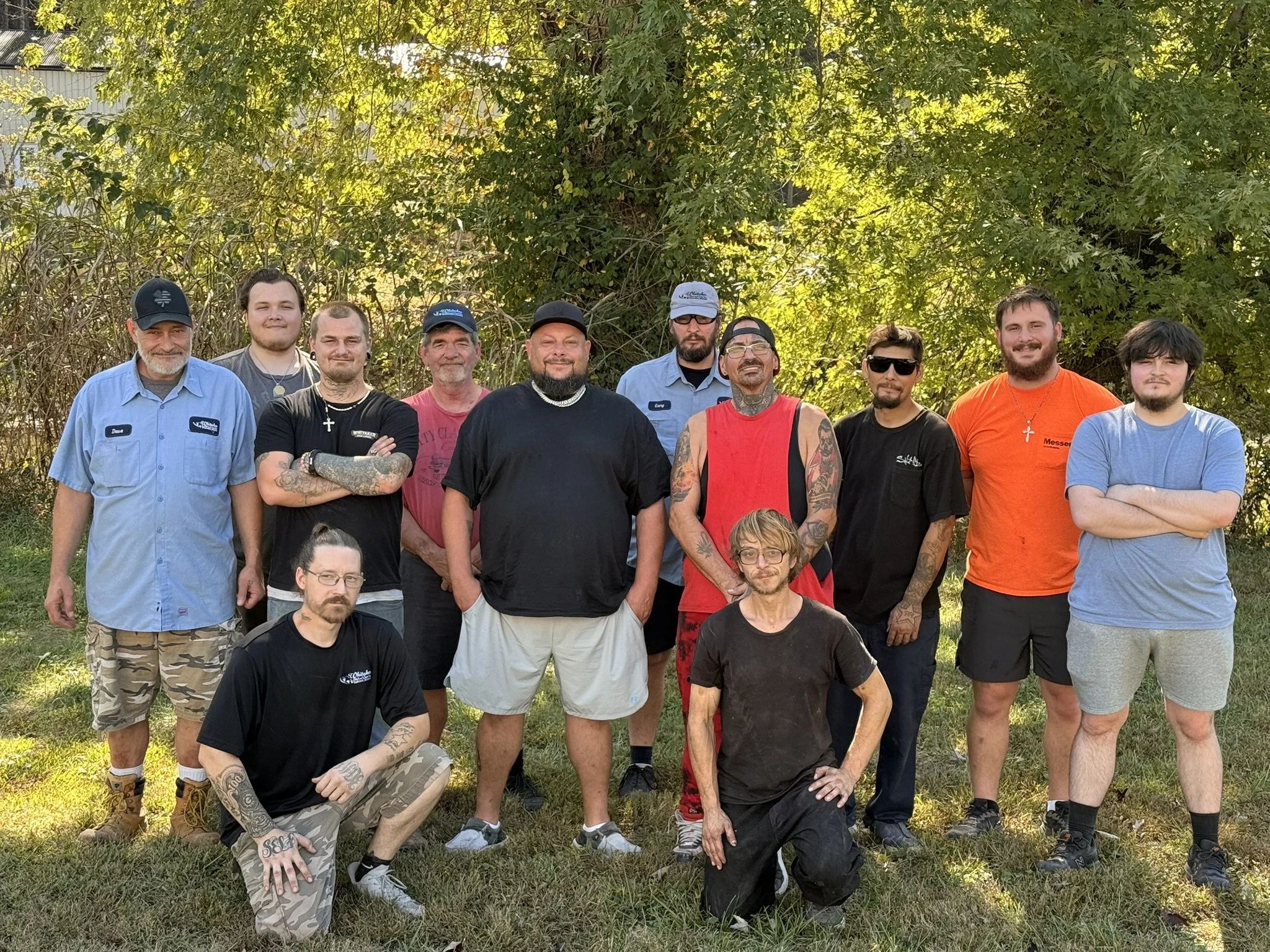 A group of twelve people standing outdoors in front of greenery, some smiling and looking at the camera, with a mix of casual clothing and tattoos visible.