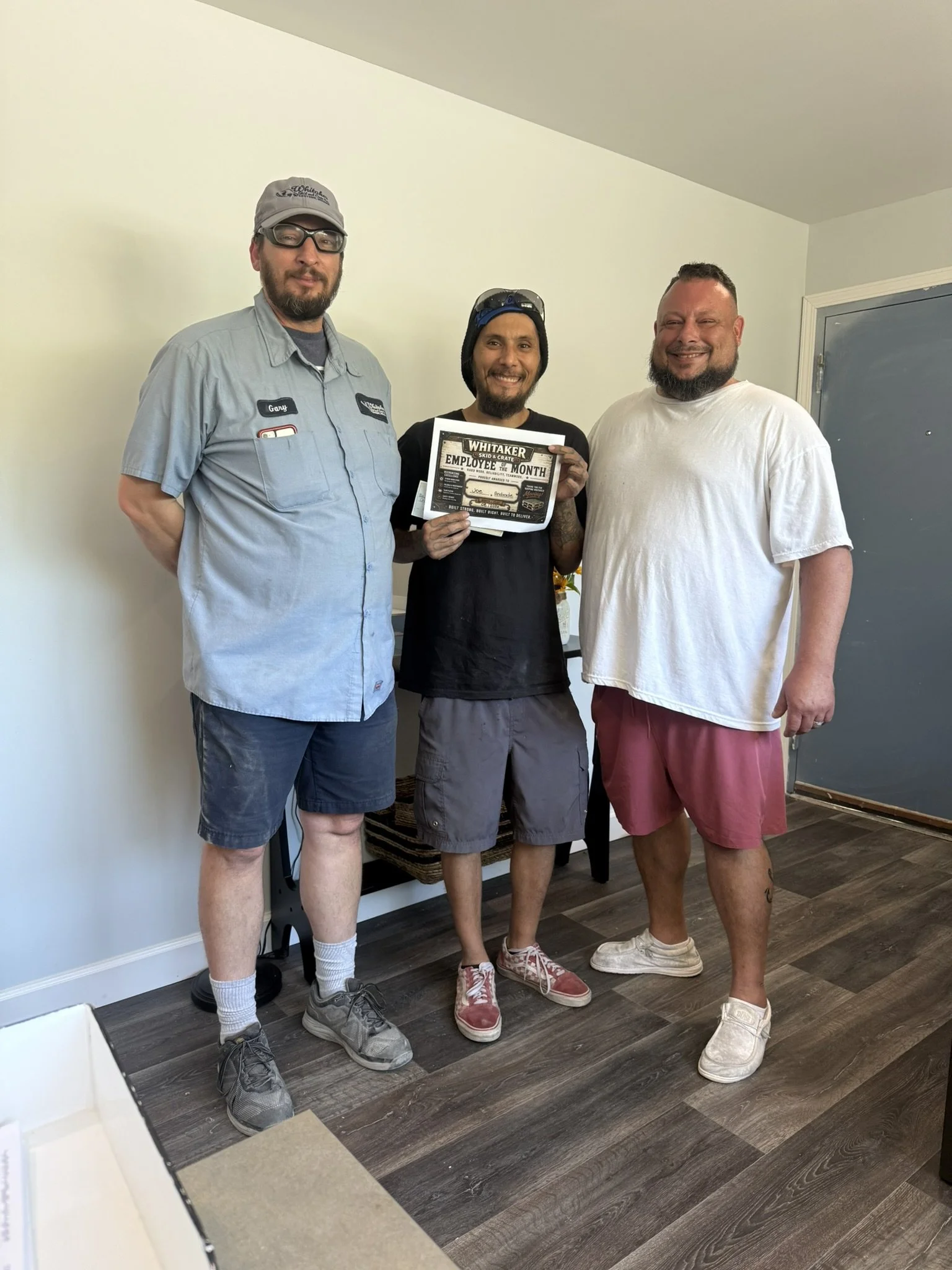 Three men standing indoors, one holding a framed certificate titled "Employee of the Month" from Whitaker, smiling for the photo.