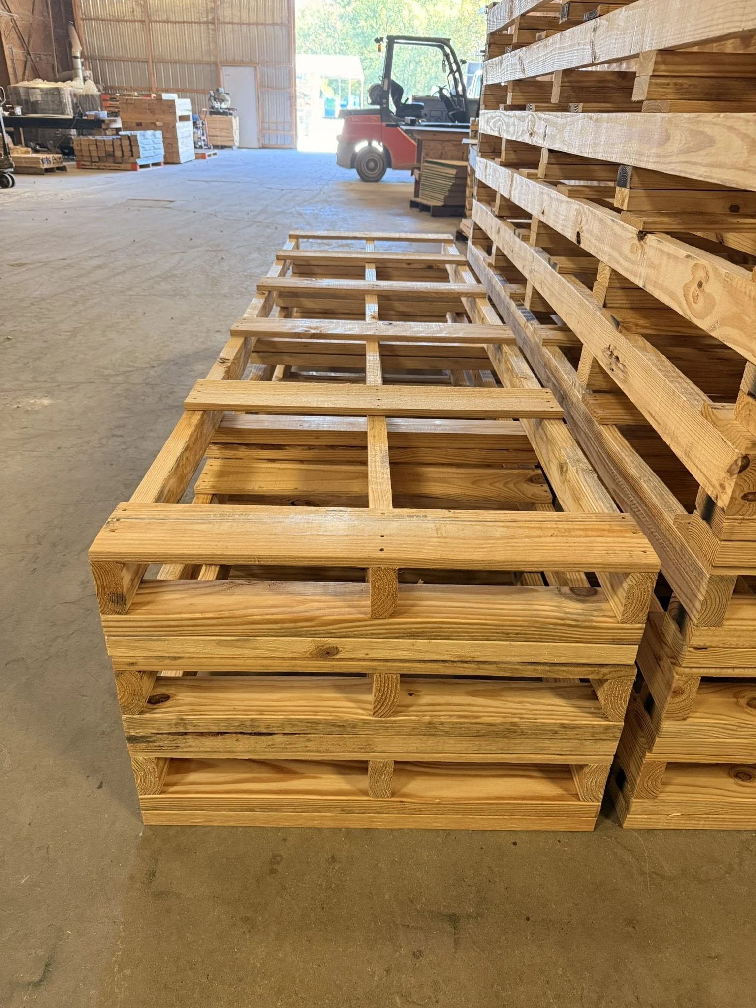 A large wooden pallet in the foreground and stacked pallets in the background inside a warehouse with a forklift and workbenches.