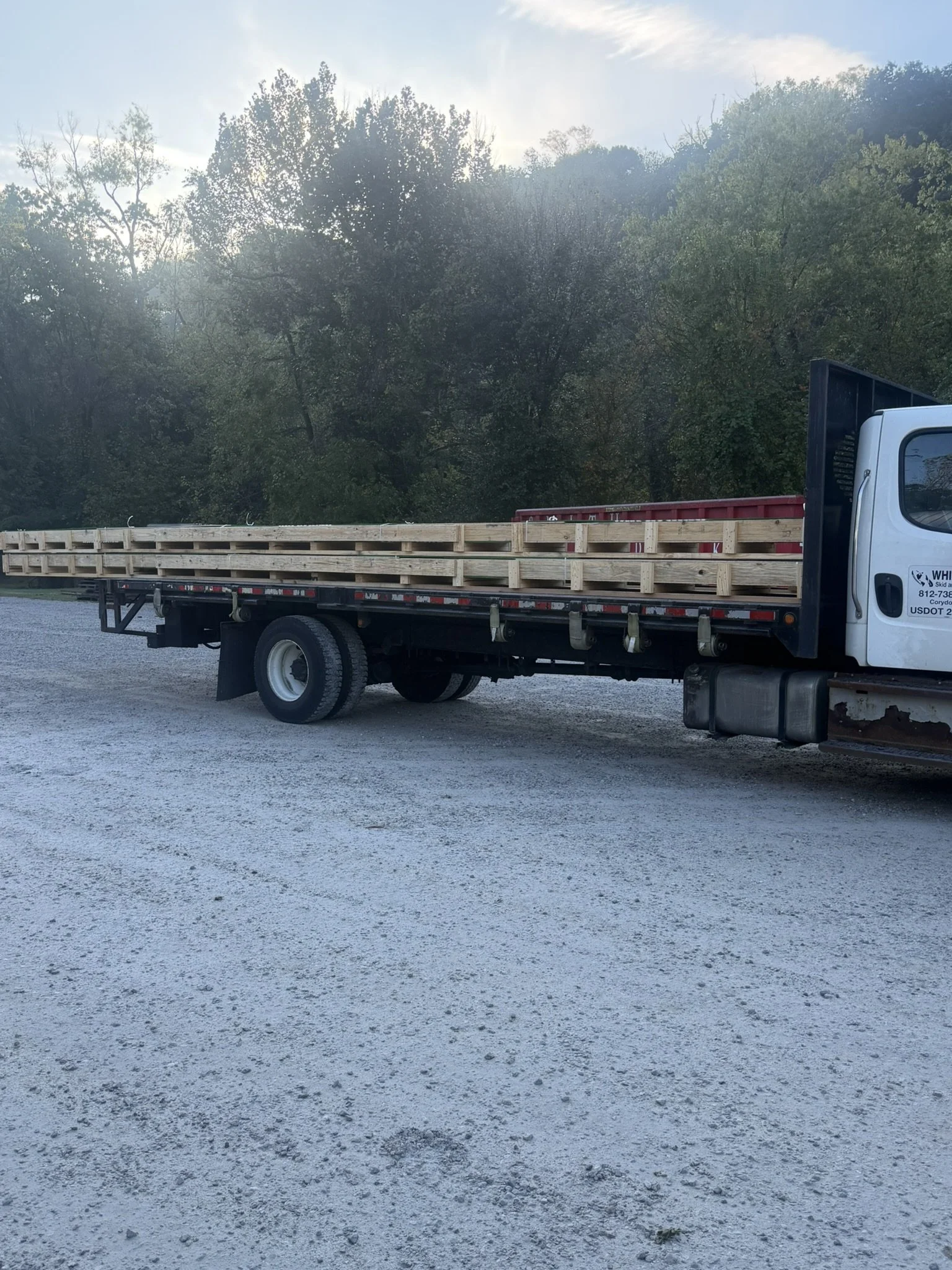 A flatbed truck carrying wooden pallets and red equipment parked on a gravel surface with a background of trees and a partly cloudy sky.