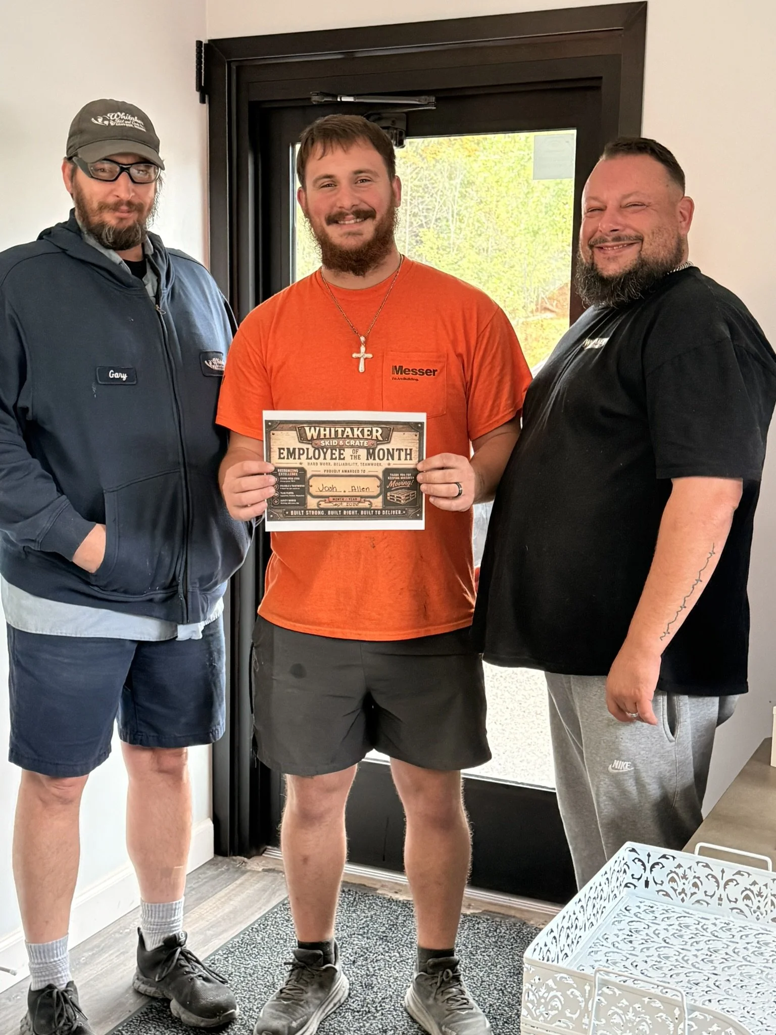 Three men standing inside a house by a door, smiling at the camera. The man in the middle is holding a plaque that reads 'Employee of the Month.' The man on the left is wearing a baseball cap, glasses, a navy jacket, and shorts. The man on the right is wearing a black shirt and gray sweatpants. The man in the middle has a beard, mustache, and is wearing an orange shirt with a cross necklace.
