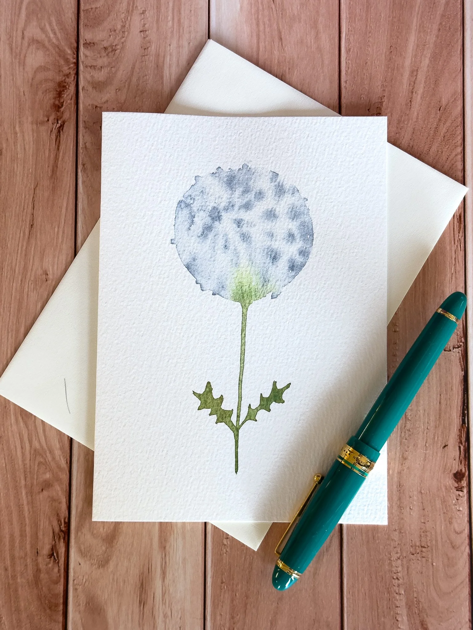Watercolor painting of a soft grey dandelion flower with green leaves on a textured white paper, placed on a wooden surface with a teal pen nearby.