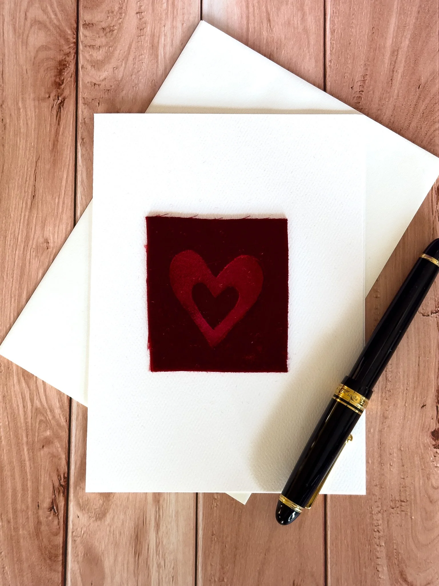 A white greeting card with a red embossed velvet heart pattern on the front, placed on a white envelope and a wooden surface, next to a black and gold pen.