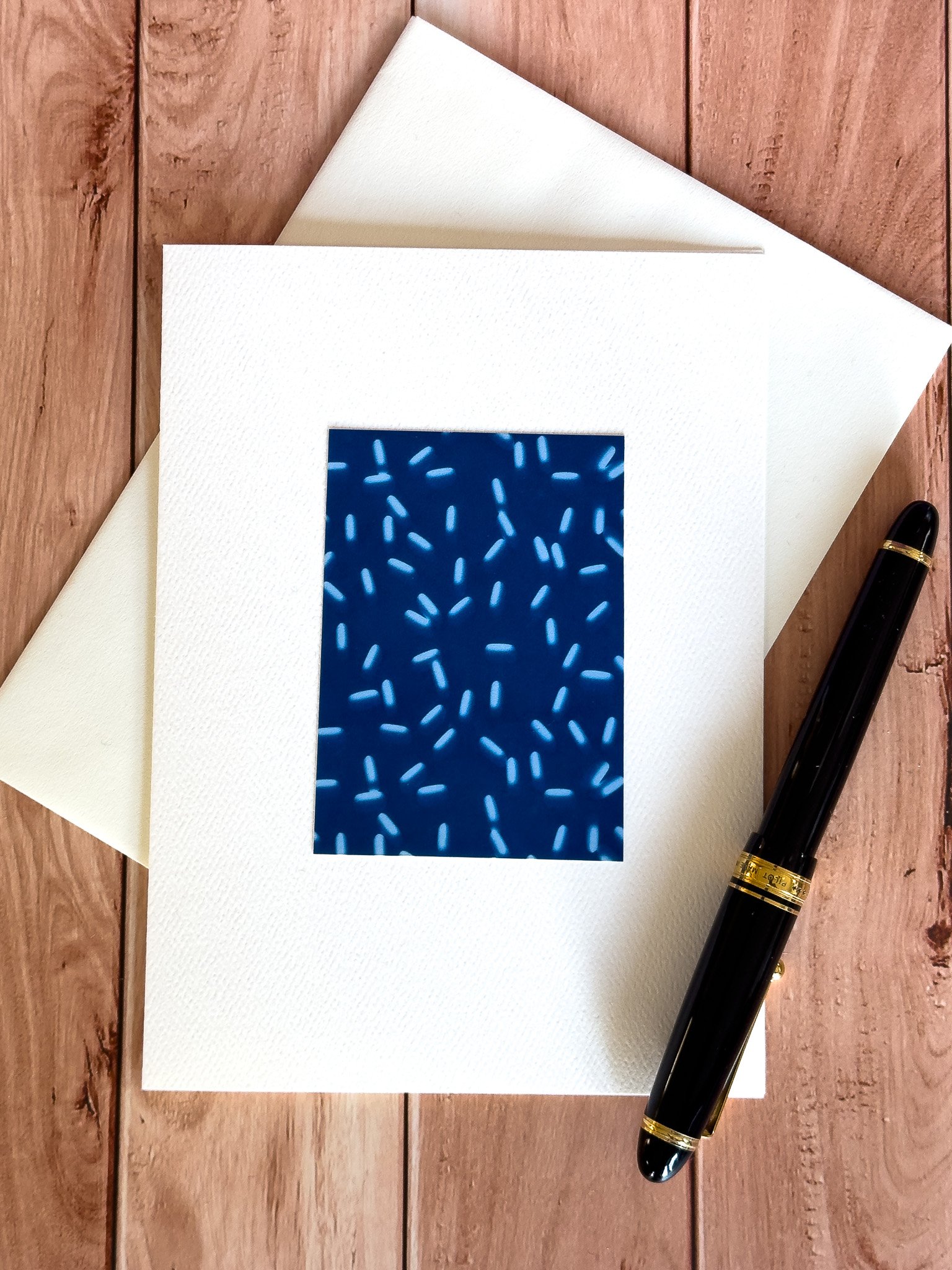A white greeting card with a cyanotype print of a small white pills, placed on a wooden surface with a black pen nearby.