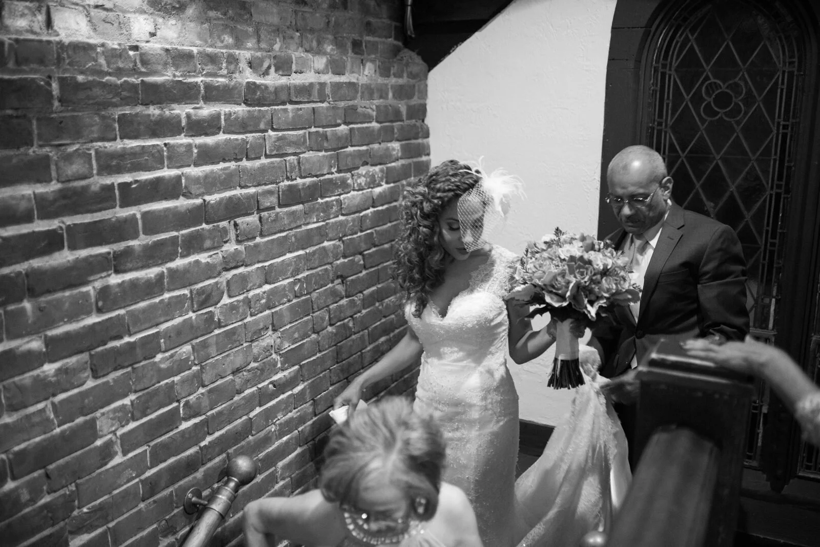 Black-and-white photo of a bride and her father walking down a brick-walled staircase on the wedding day
