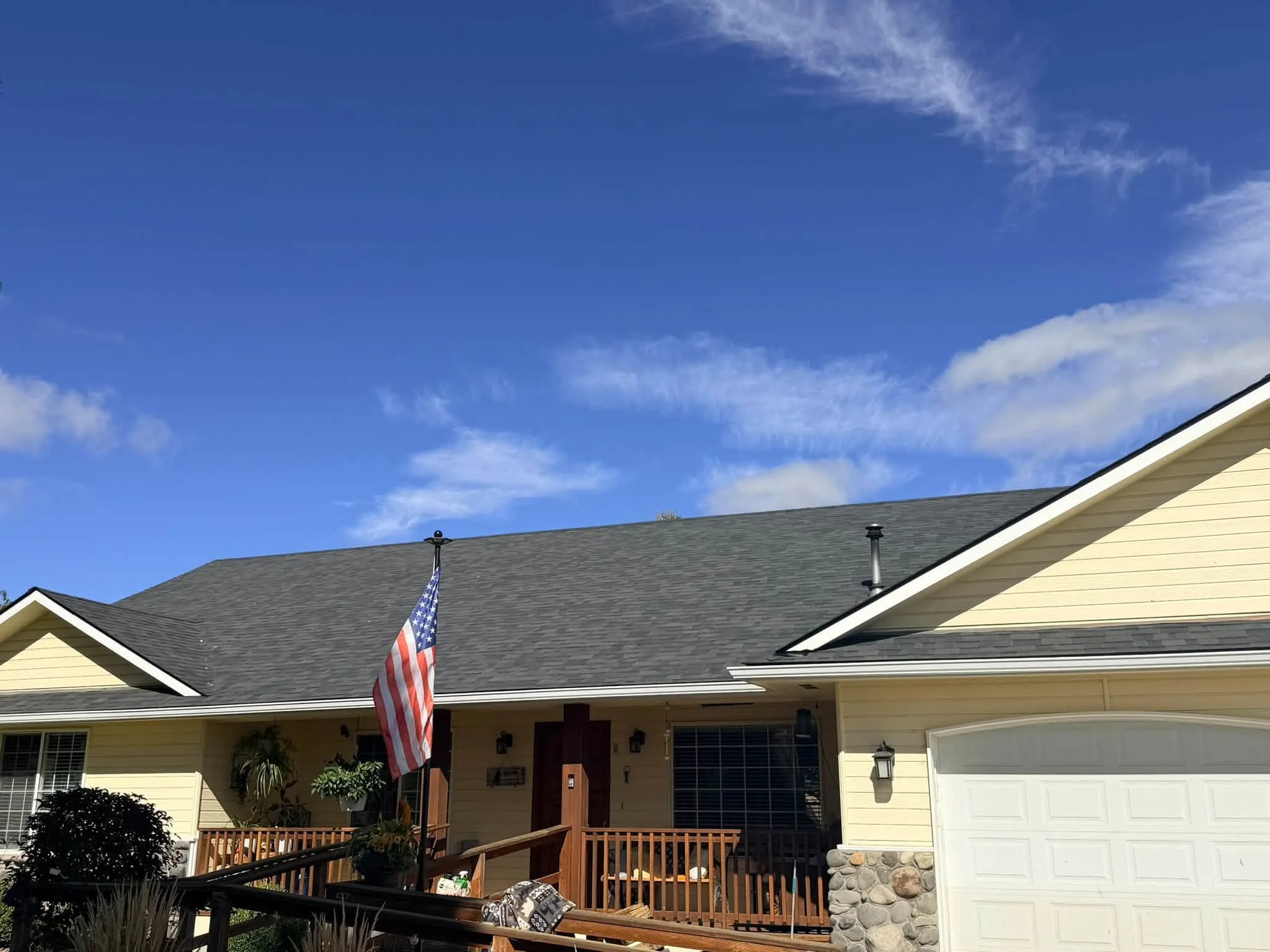 A yellow house with a gray shingled roof and a front porch, with an American flag hanging in the yard, under a blue sky with some scattered clouds.
