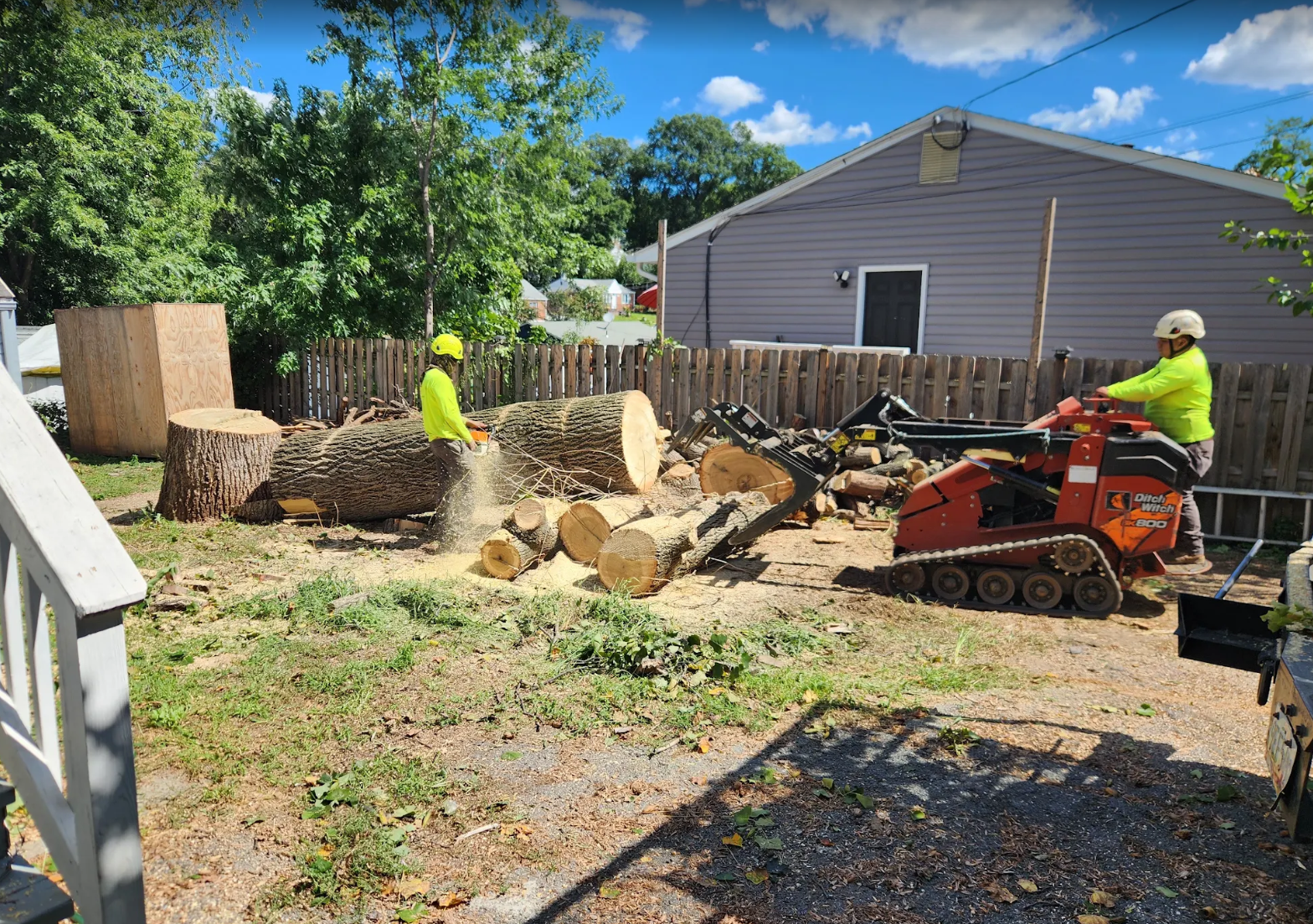 Two workers in bright yellow safety gear and helmets cut and move large fallen tree logs in a backyard with a wood chipper machine.