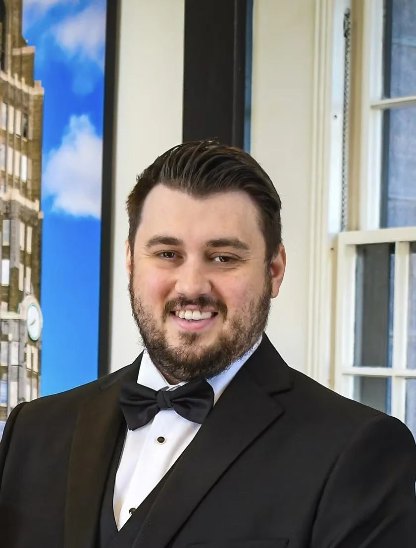 A man in a black tuxedo with a bow tie, smiling, standing indoors near a window with a cityscape and blue sky visible outside.