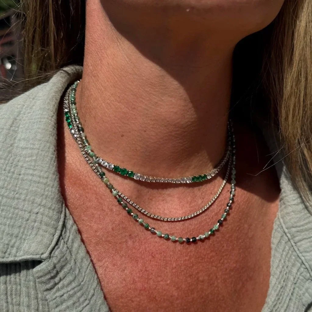 A close-up of a person's neck and collarbone area, wearing three layered necklaces, with part of a light-colored shirt visible.