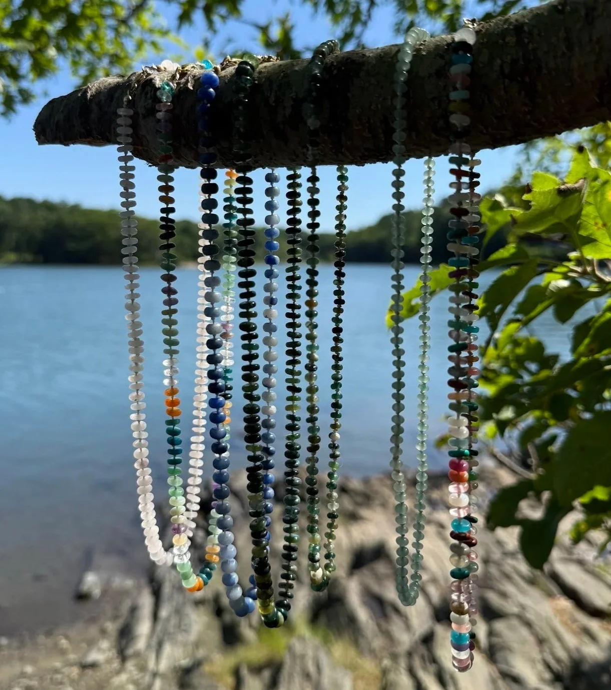 Colorful beaded necklaces hanging from a tree branch near a lake, with green leaves and a clear blue sky in the background.