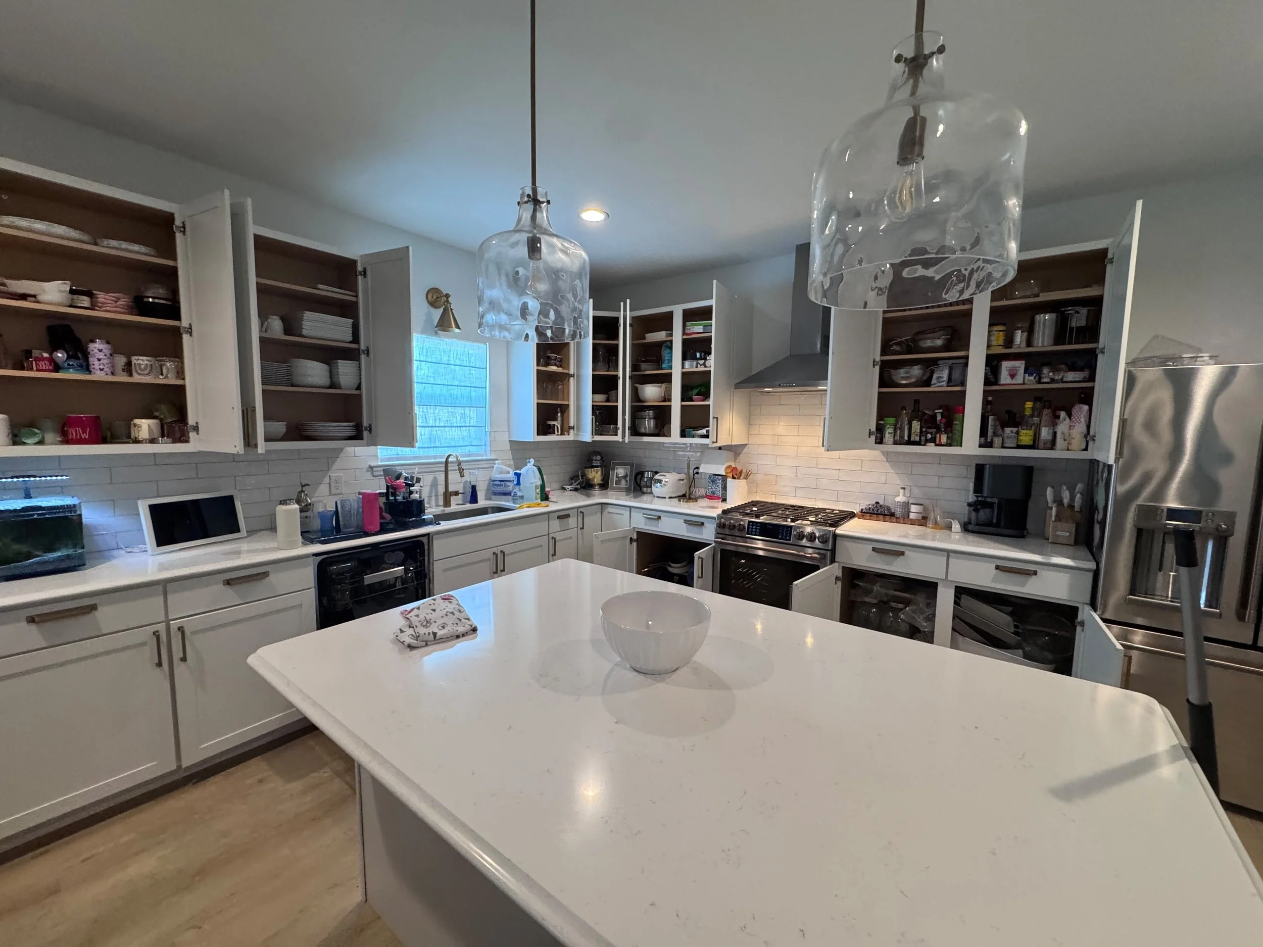 A modern kitchen with white cabinets, open shelves, and a large kitchen island with a white countertop. There are two glass pendant lights hanging from the ceiling and various kitchen appliances and utensils on the counter. A small bowl and a towel a