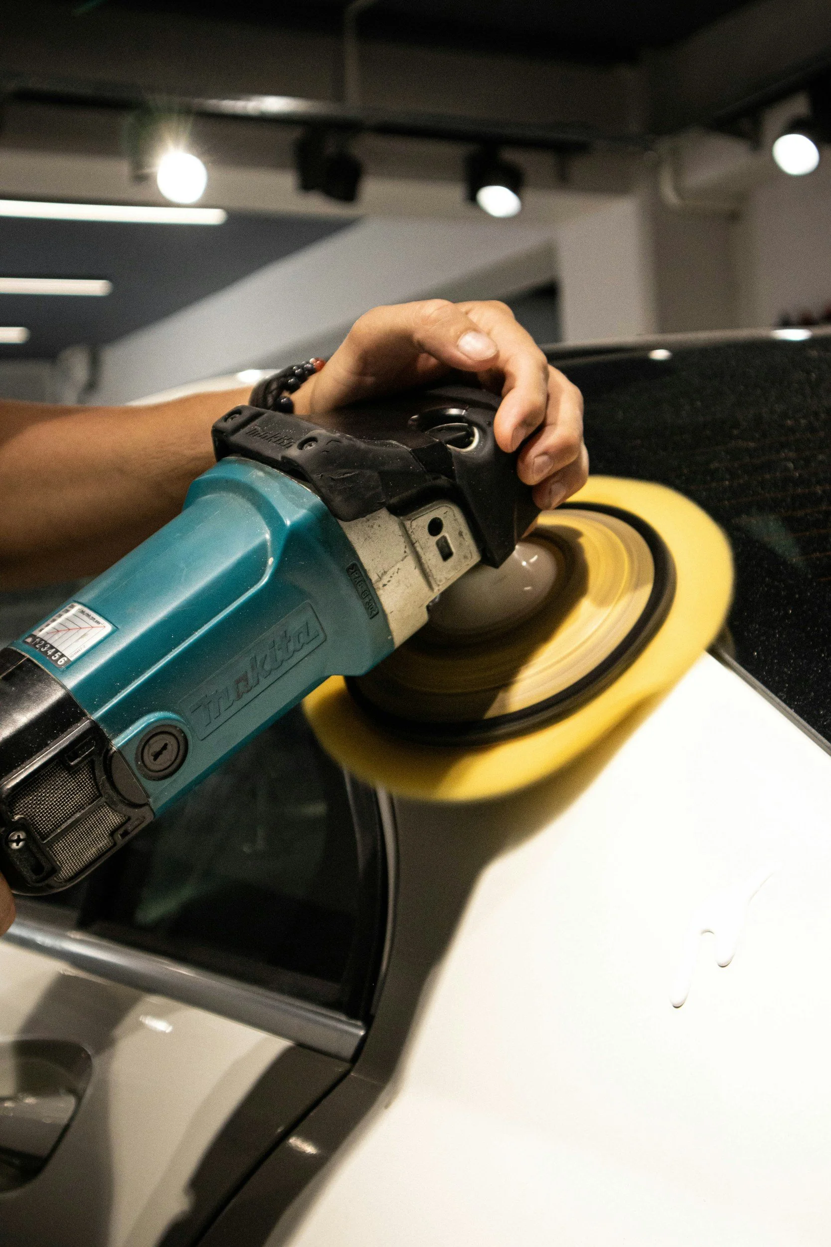 Close-up of a person polishing a black car with a power buffer using a yellow polishing pad in a garage or workshop.