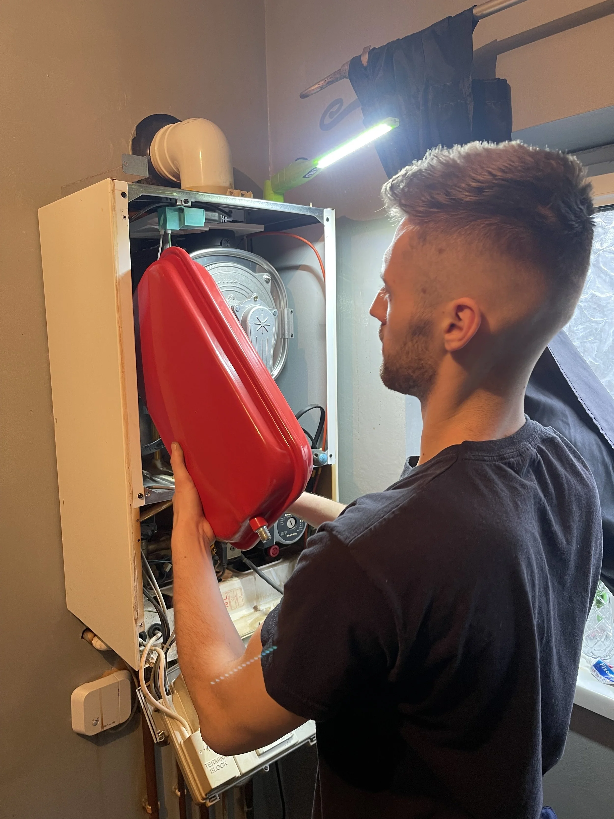 A man working on a combi boiler, holding a red expansion vessel, with a flashlight illuminating the panel.