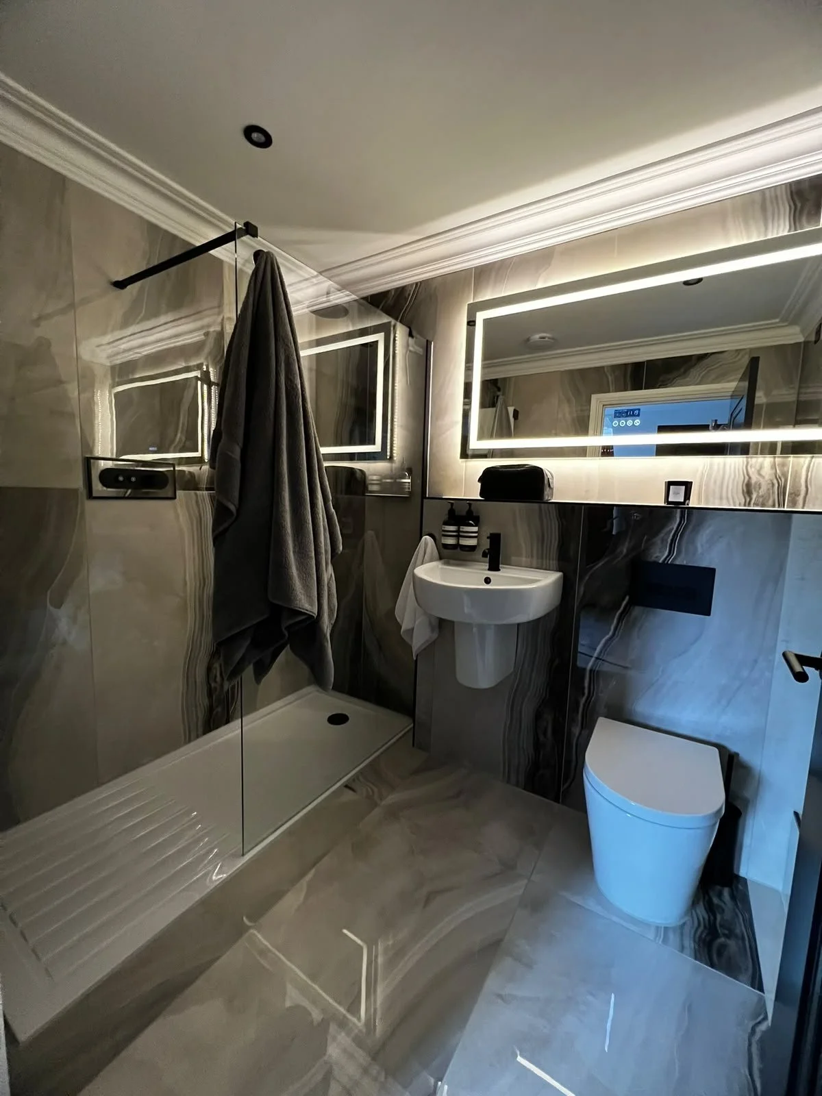 Modern bathroom with beige marble walls, a walk-in shower with glass door, towel hanging on shower rod, illuminated rectangular mirror above sink, white sink with black faucet, black toiletries bottles, wall-mounted toilet, and a small window with a digital clock visible outside.