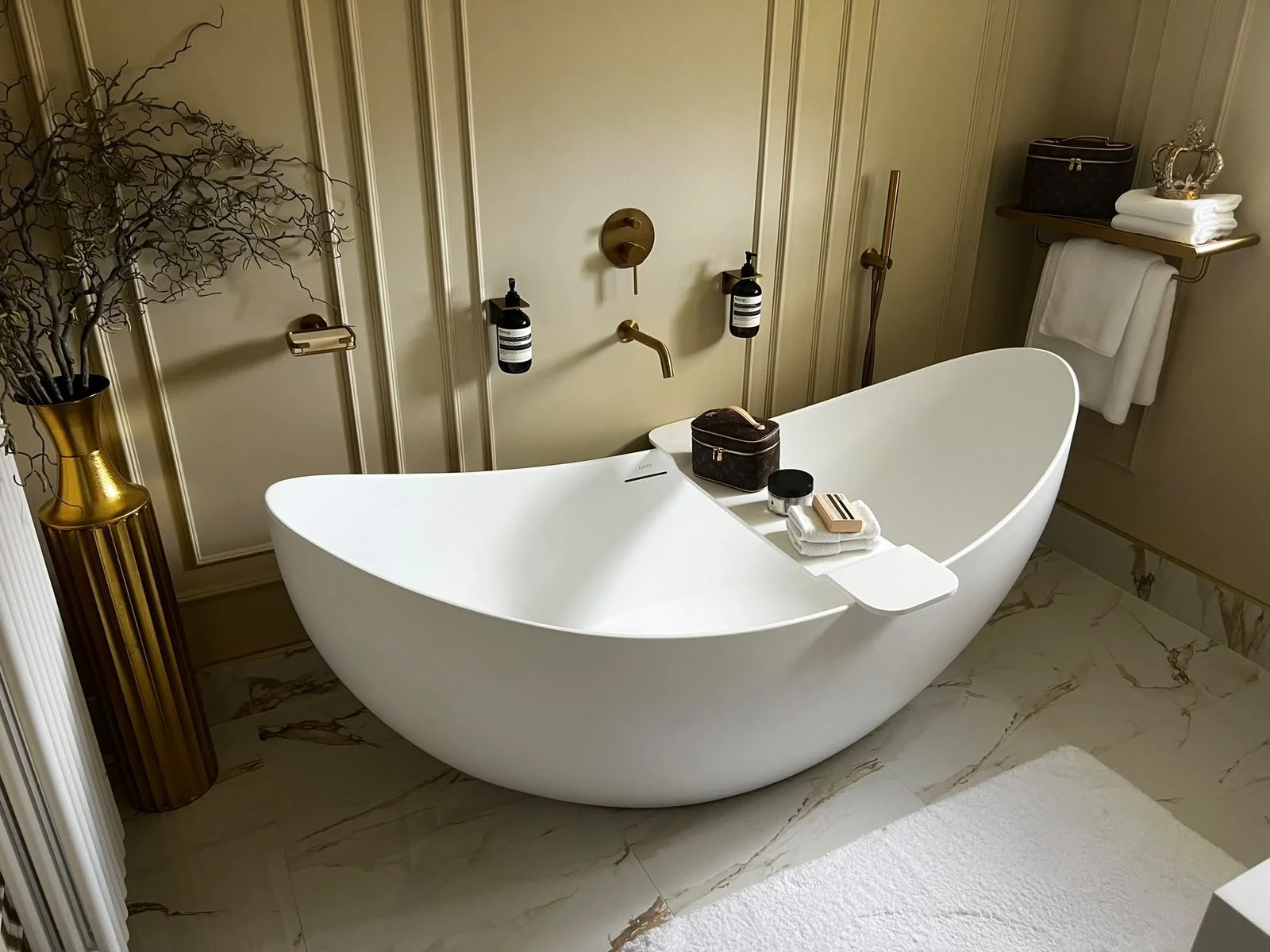 Luxurious bathroom with a modern freestanding bathtub, gold accents, and elegant decor, including a tall gold vase with decorative branches, neatly folded towels, and personal care products on the bathtub ledge.