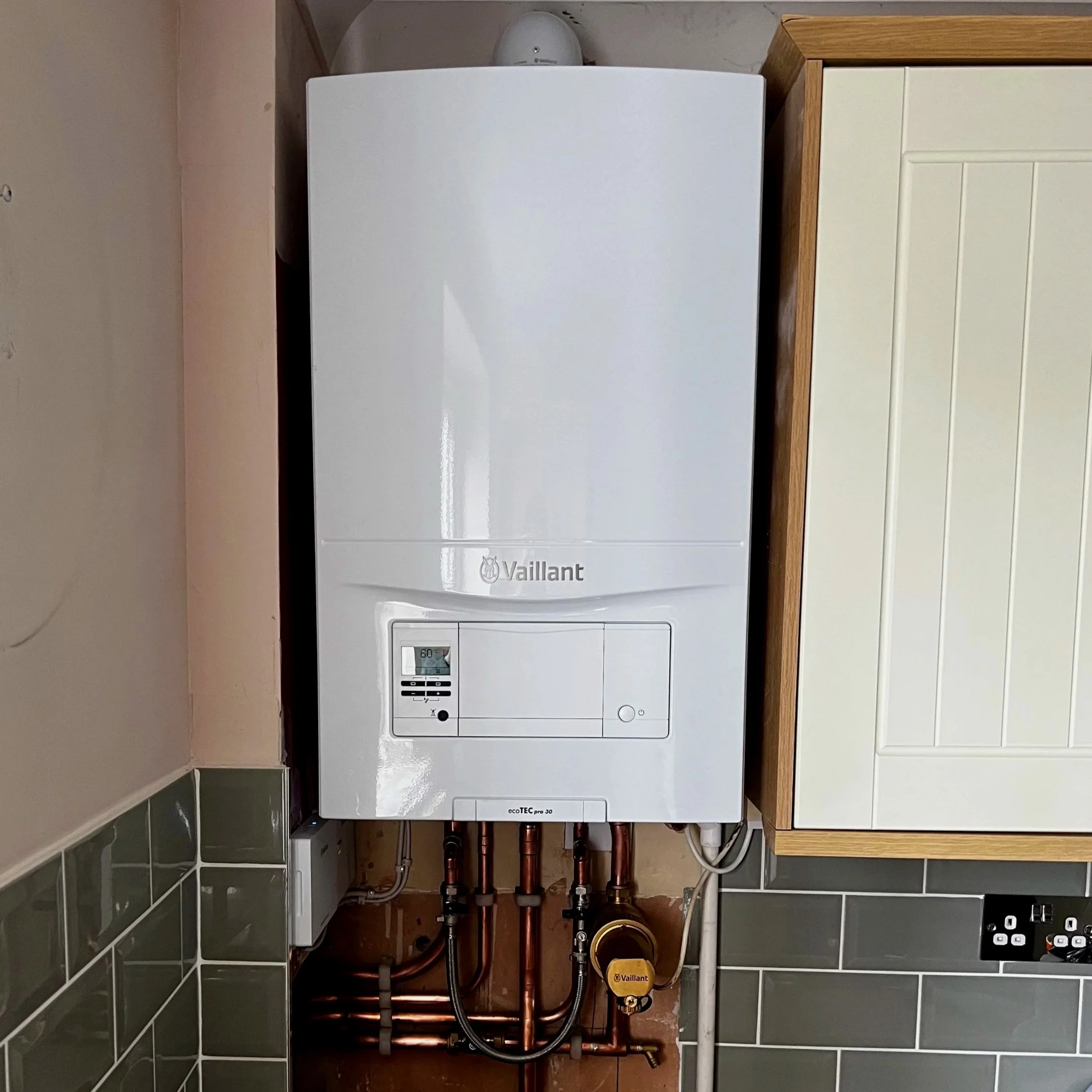 White Vaillant ecoTEC pro 30 boiler mounted on a wall above copper pipes and a yellow valve, with green and white tiled wall below and beige wall to the side.