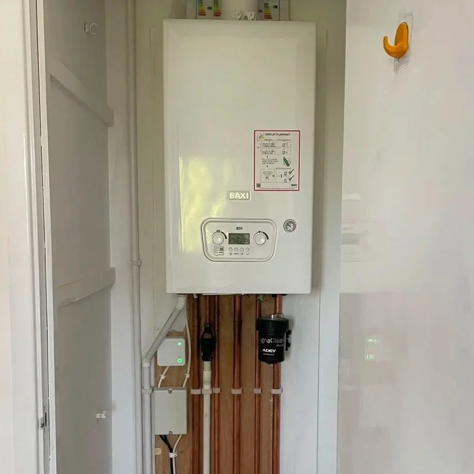 A wall-mounted Baxi boiler with control panel, copper pipes, and a water filter attached below, located in a utility or laundry room.
