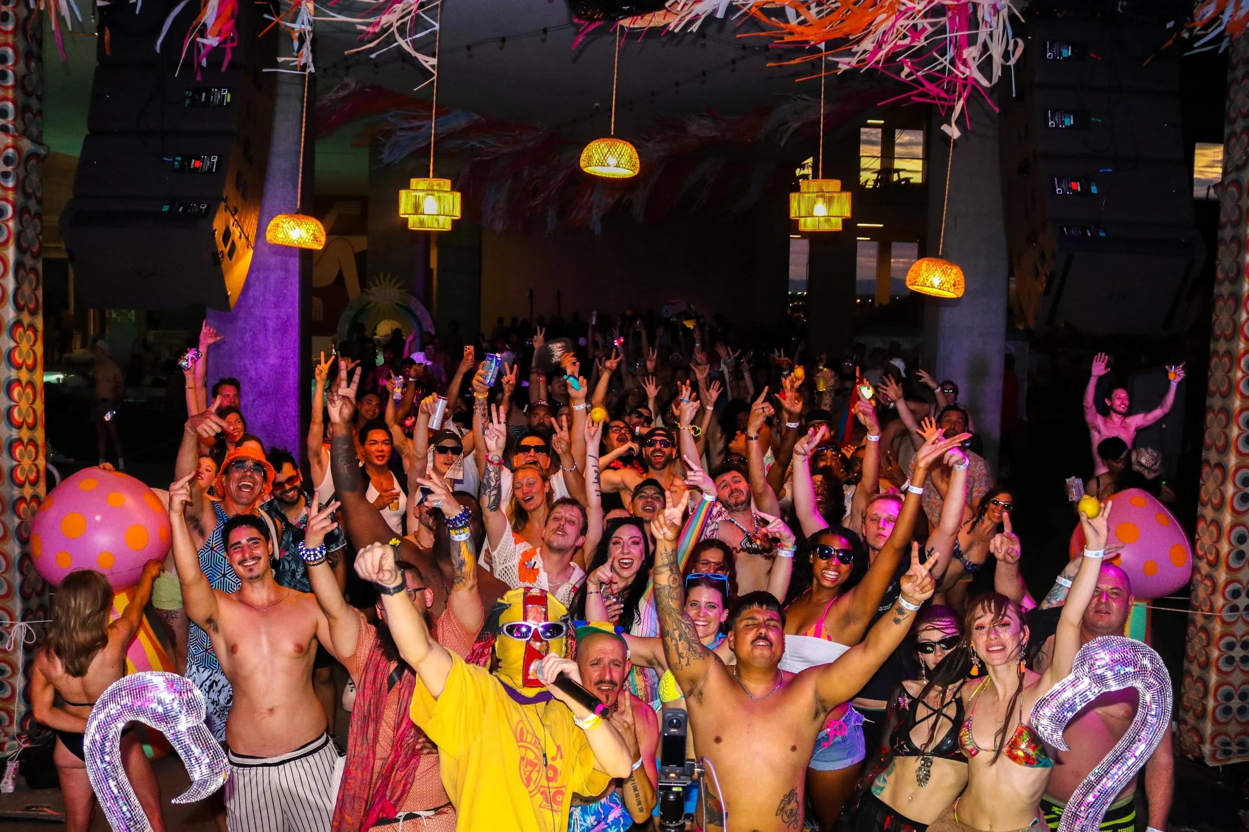 A large crowd of people at a lively party or concert, many wearing colorful outfits and accessories, some with tattoos and sunglasses. The atmosphere is vibrant with hanging lights and decorations, and people are enjoying themselves with raised hands and smiling faces.