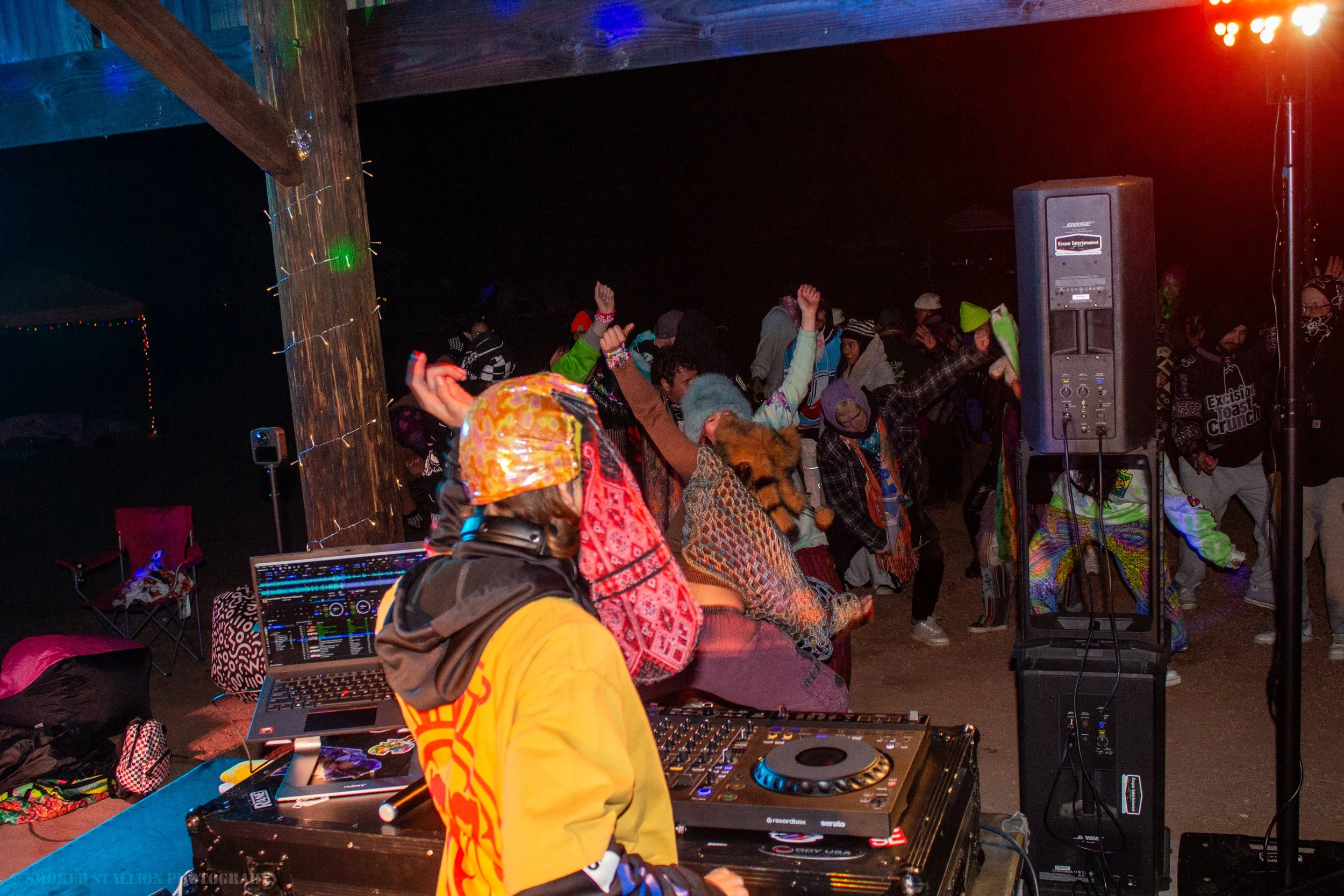 A DJ wearing a yellow jacket and a colorful hat is playing music at an outdoor nighttime event. People are dancing in front of the DJ, with colorful lights and decorations visible in the background.