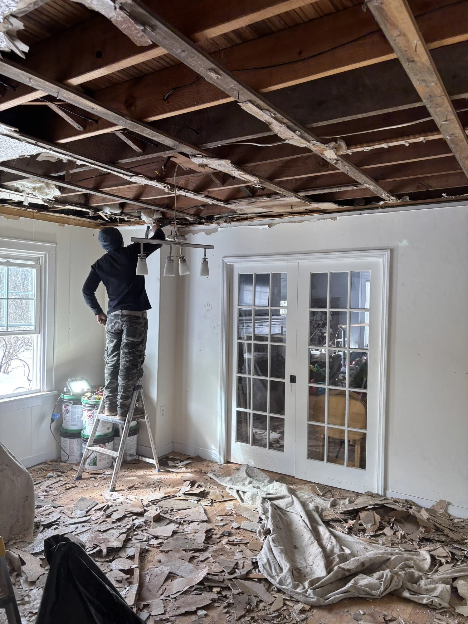 A person standing on a ladder, removing ceiling drywall in a room under renovation, with debris on the floor and materials nearby.