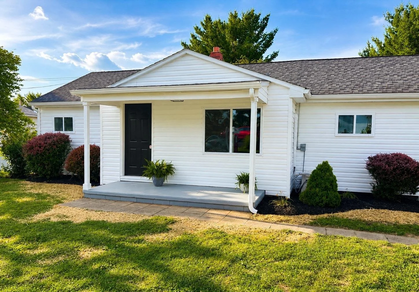 A white house with a small front porch, black door, and two potted plants. The house has a gabled roof, two windows, and is surrounded by shrubs, trees, and a grassy lawn under a partly cloudy sky.