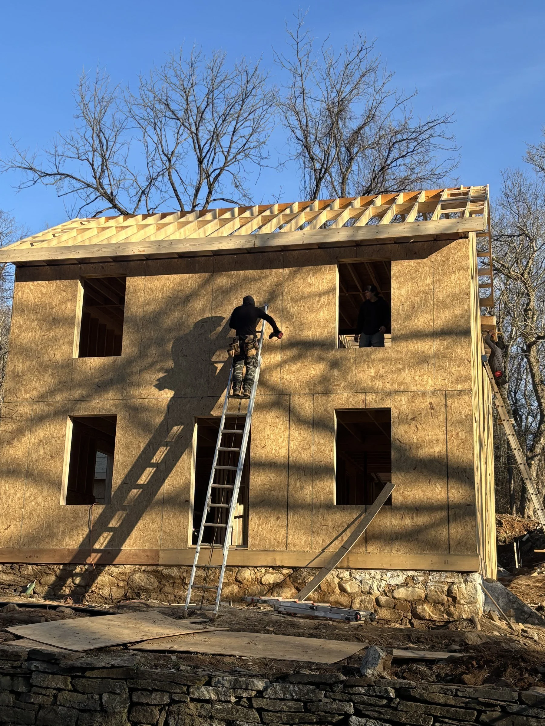 Construction workers building the wooden frame of a house, ladders leaning against the structure, with trees and a blue sky in the background.