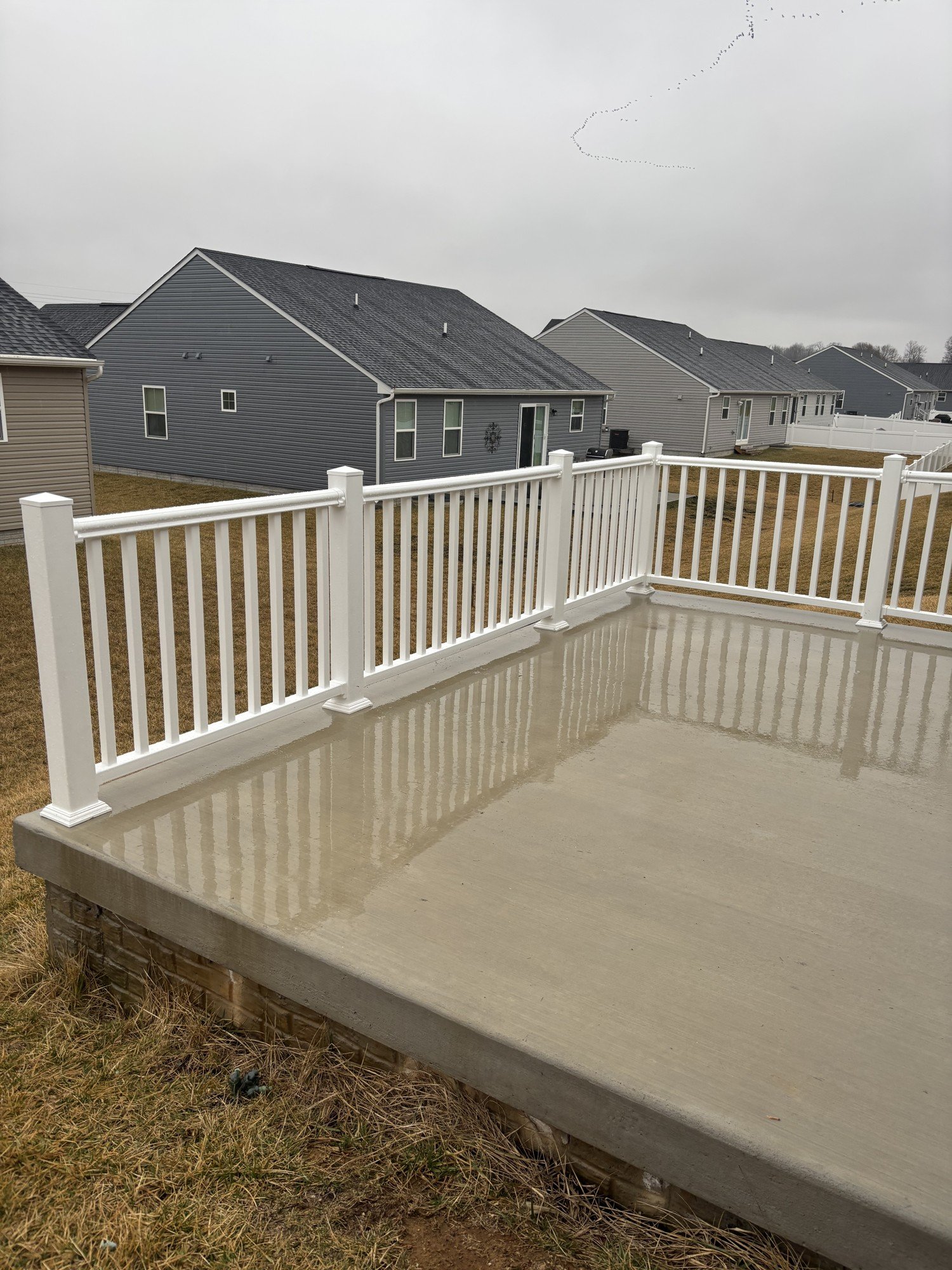 View of a backyard deck with a white railing and wet concrete surface, with neighboring houses and a cloudy sky visible in the background.