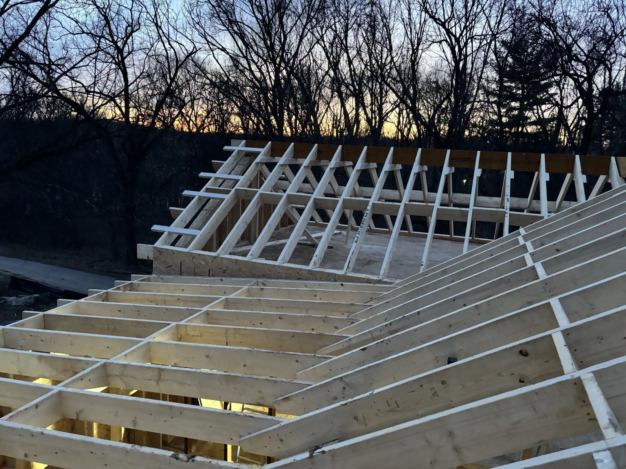 Construction site with wooden framing of a building under construction, set against a background of leafless trees and a sunset sky.