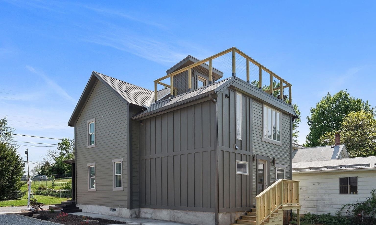 Back view of a modern gray two-story house with a rooftop deck enclosed by a wooden railing, and a small staircase leading to the backyard, under a clear blue sky.