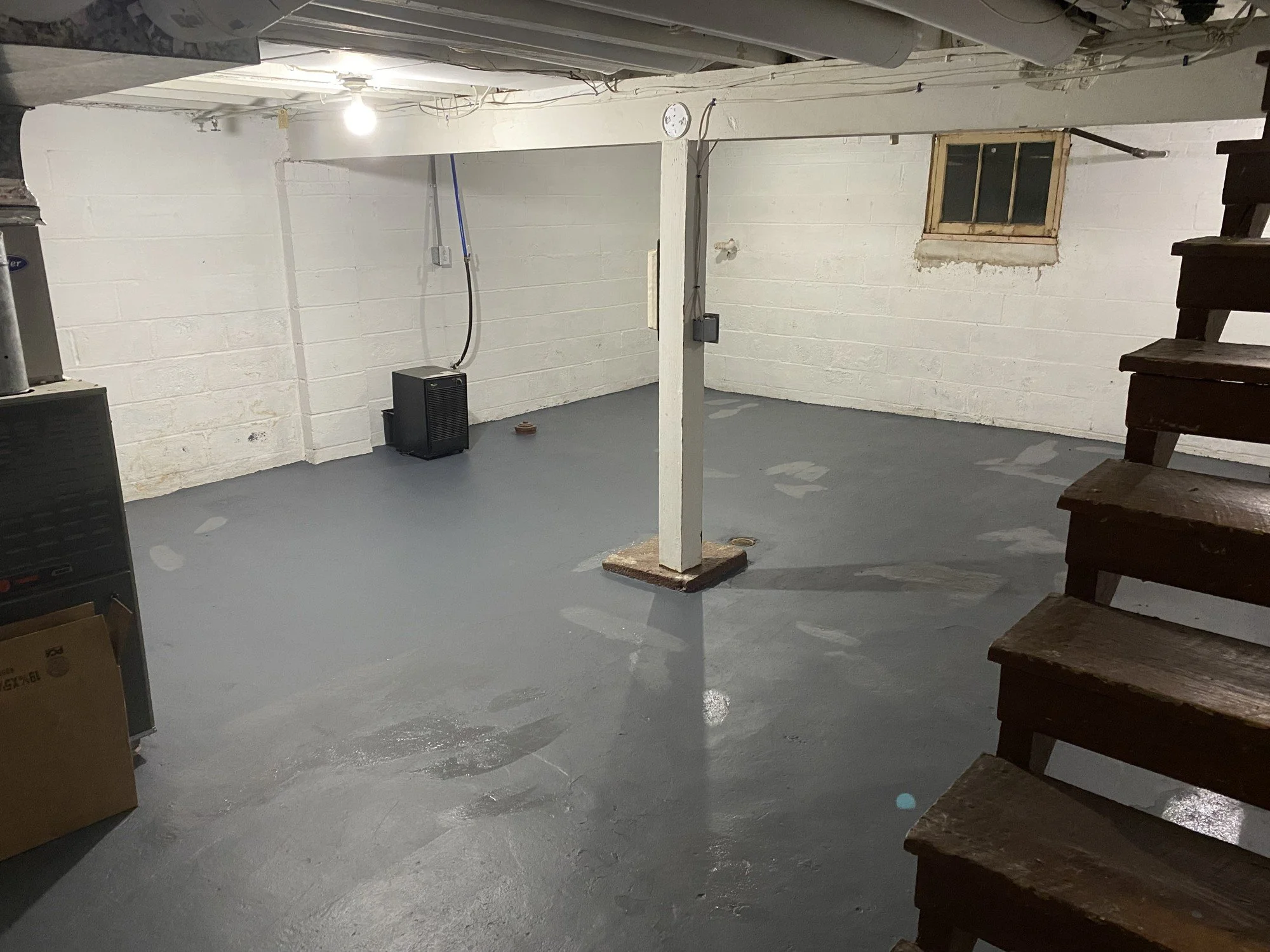 An empty basement with white painted brick walls, a gray concrete floor, a wooden staircase on the right, a small window on the wall, and a single light bulb hanging from the ceiling. There is a heater in the corner and some wires and pipes on the ceiling.