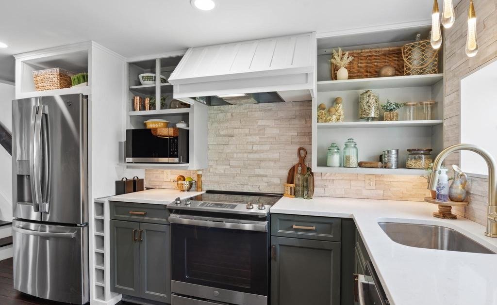Modern kitchen with stainless steel refrigerator, black oven, microwave, open shelves with decorative jars, baskets, and plants, white countertops, and a sink with gold fixtures, gray and white cabinetry, and wood accents.