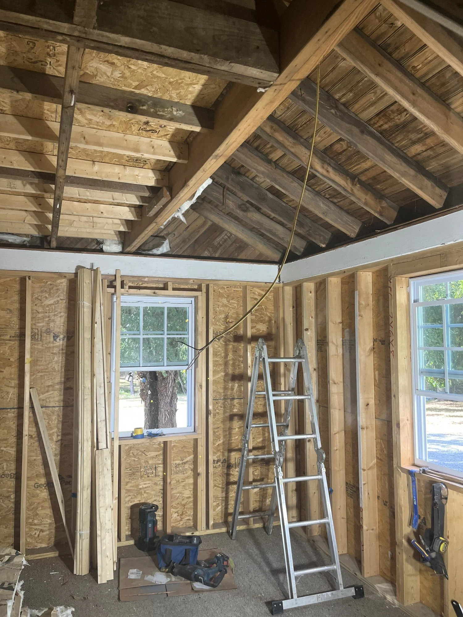 Interior of a room under construction with exposed wooden framing, tools, a step ladder, and windows providing natural light.