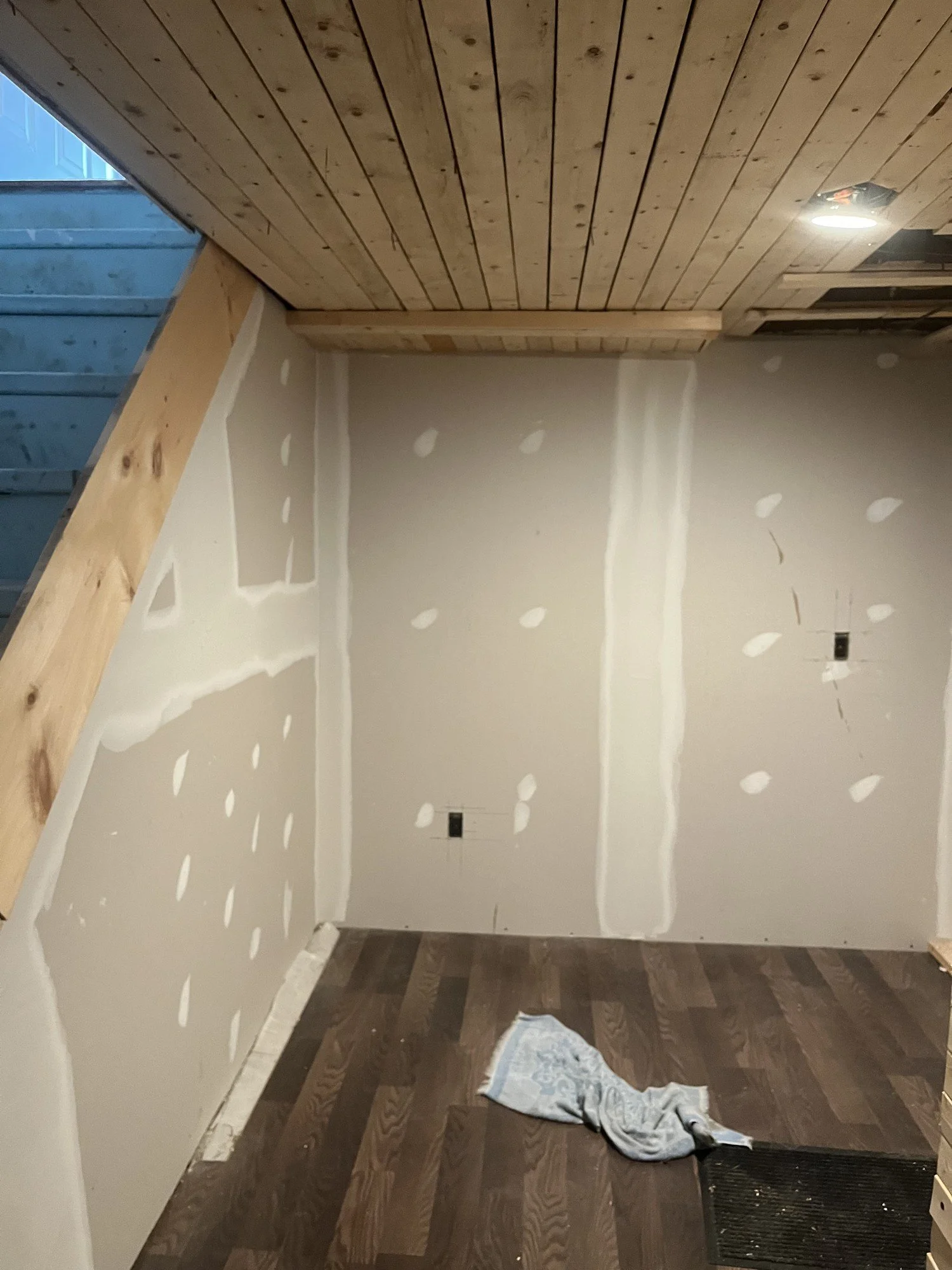 A room under construction with unfinished drywall on the walls, a heated floor, a wooden ceiling, a wooden beam on the left, and a partially installed door frame in the upper right corner. There is a piece of cloth or towel on dark brown wooden floor.
