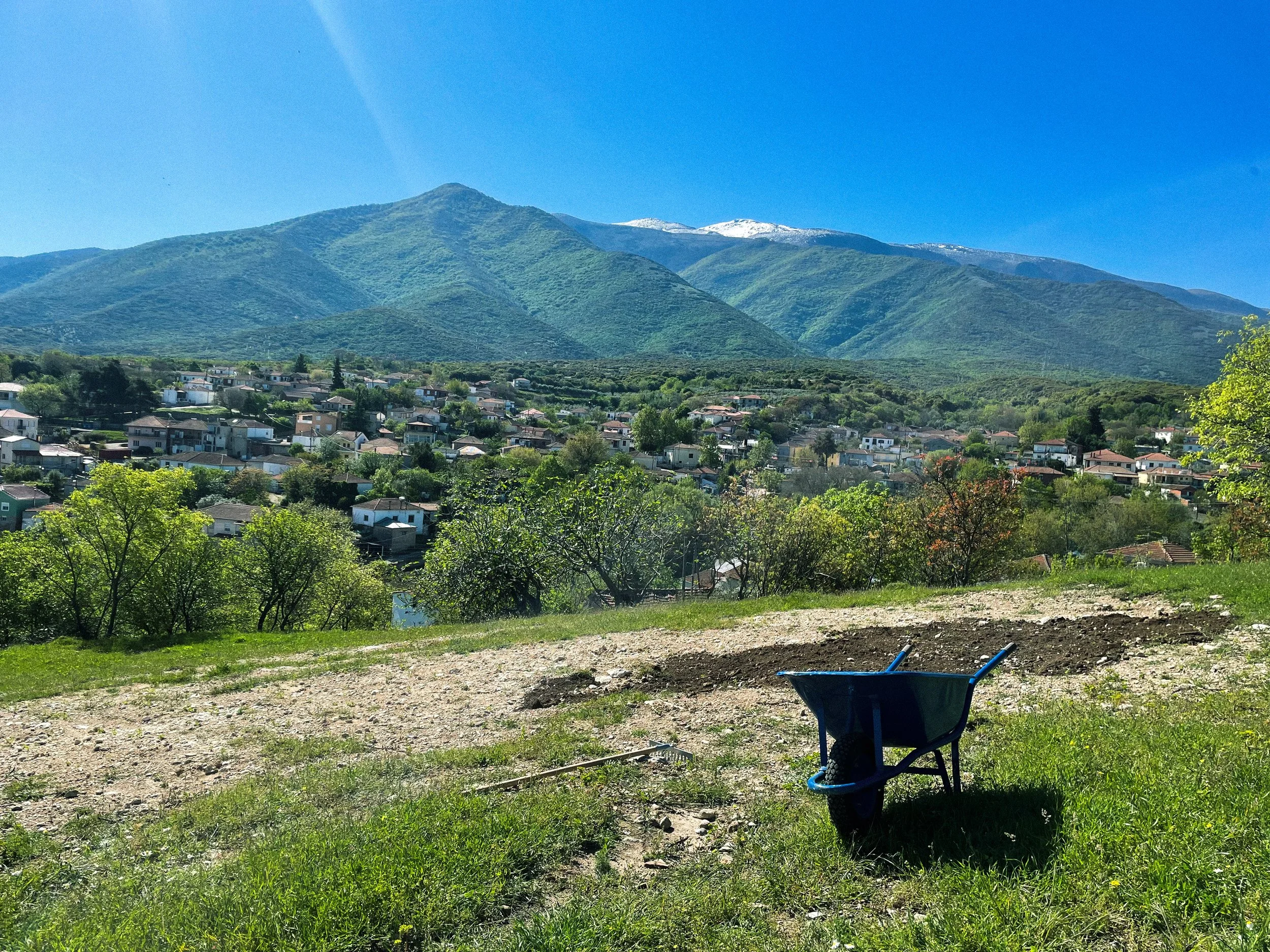 View of the Pangaion mountain with snow-capped peaks in the background, overlooking the village Kormista with houses and trees, under a clear blue sky. In the foreground, there is a wheelbarrow and some gardening tools on a patch of soil and grass.