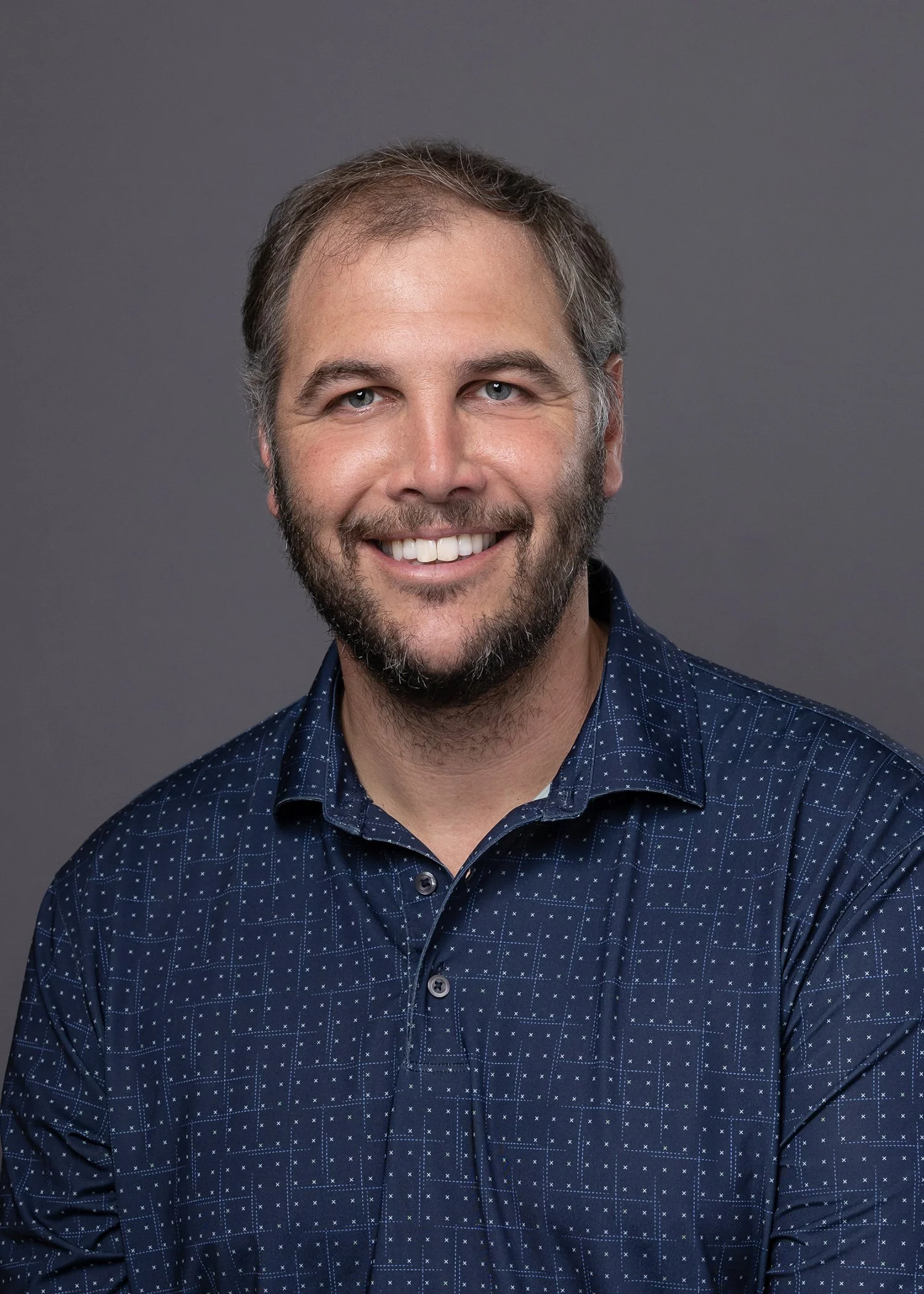 Headshot of Tom Whitwell with blue eyes, short brown hair, and a beard, wearing a navy blue patterned button-up shirt, against a plain gray background.