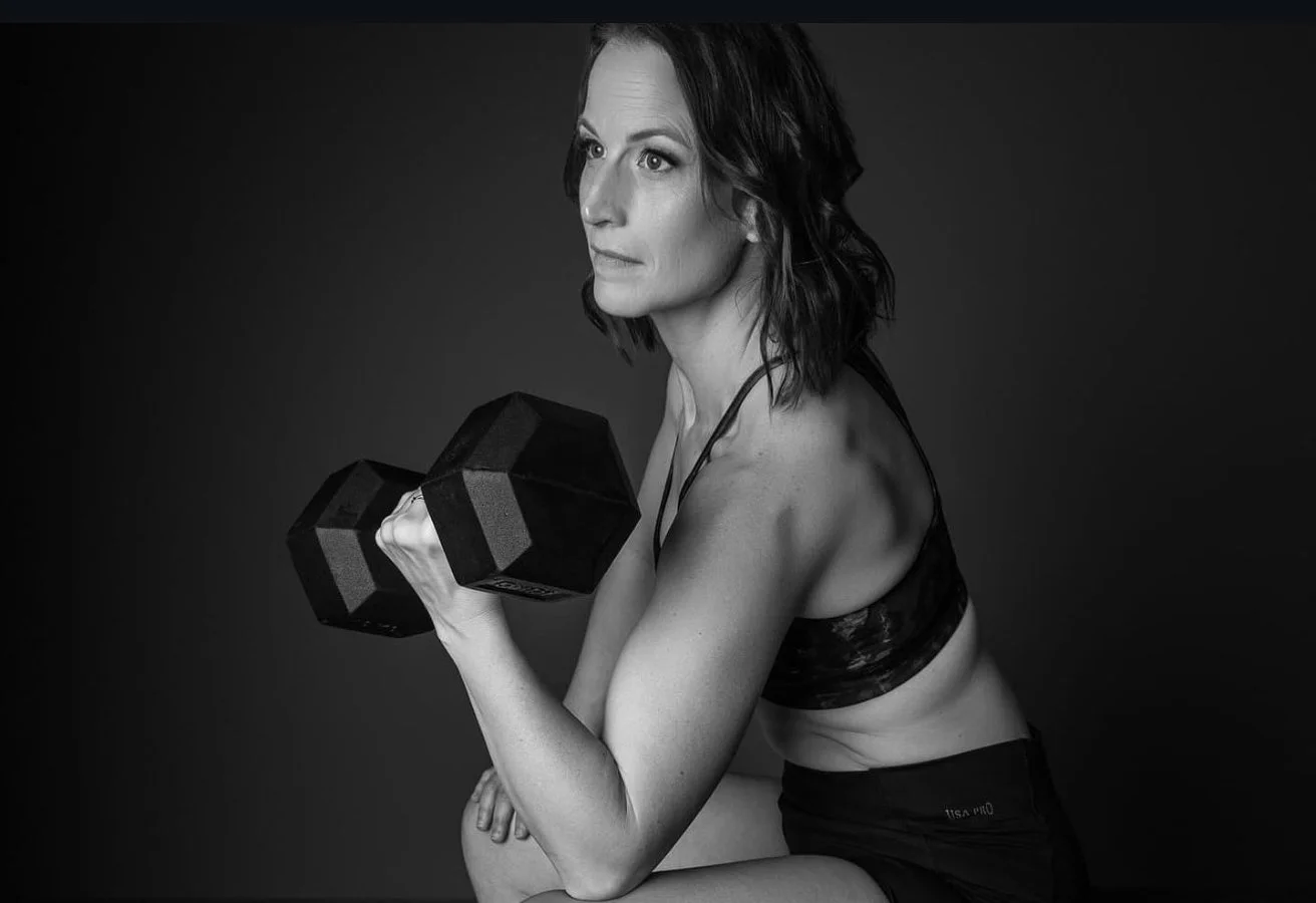 Black and white photo of a woman exercising with a dumbbell, wearing a sports bra and shorts, against a plain dark background.