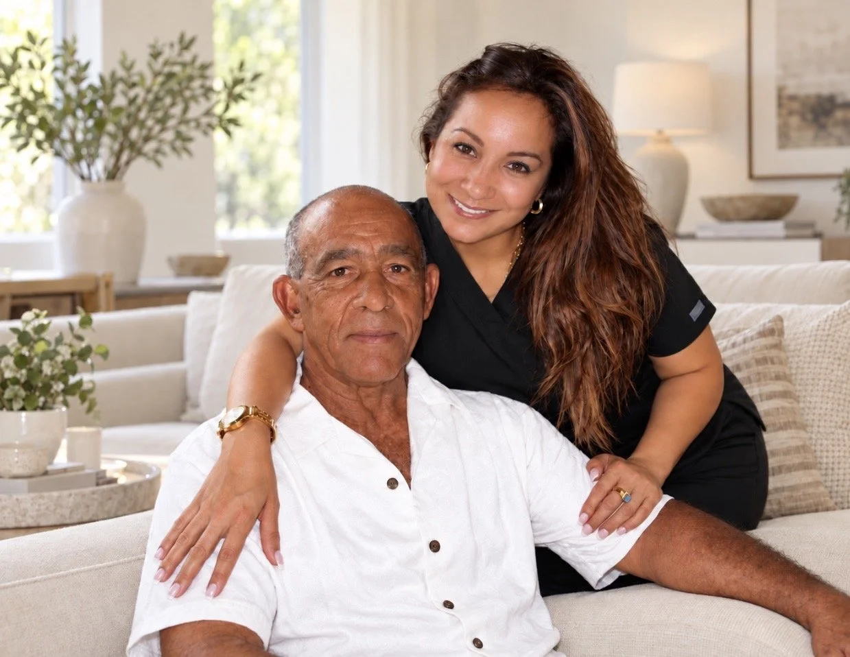 A woman with long, curly brown hair and a man with short gray hair sitting on a beige couch in a bright, well-decorated living room. The woman is smiling and wrapping her arms around the man's shoulders. The man is wearing a white button-up shirt and the woman is dressed in black scrubs.