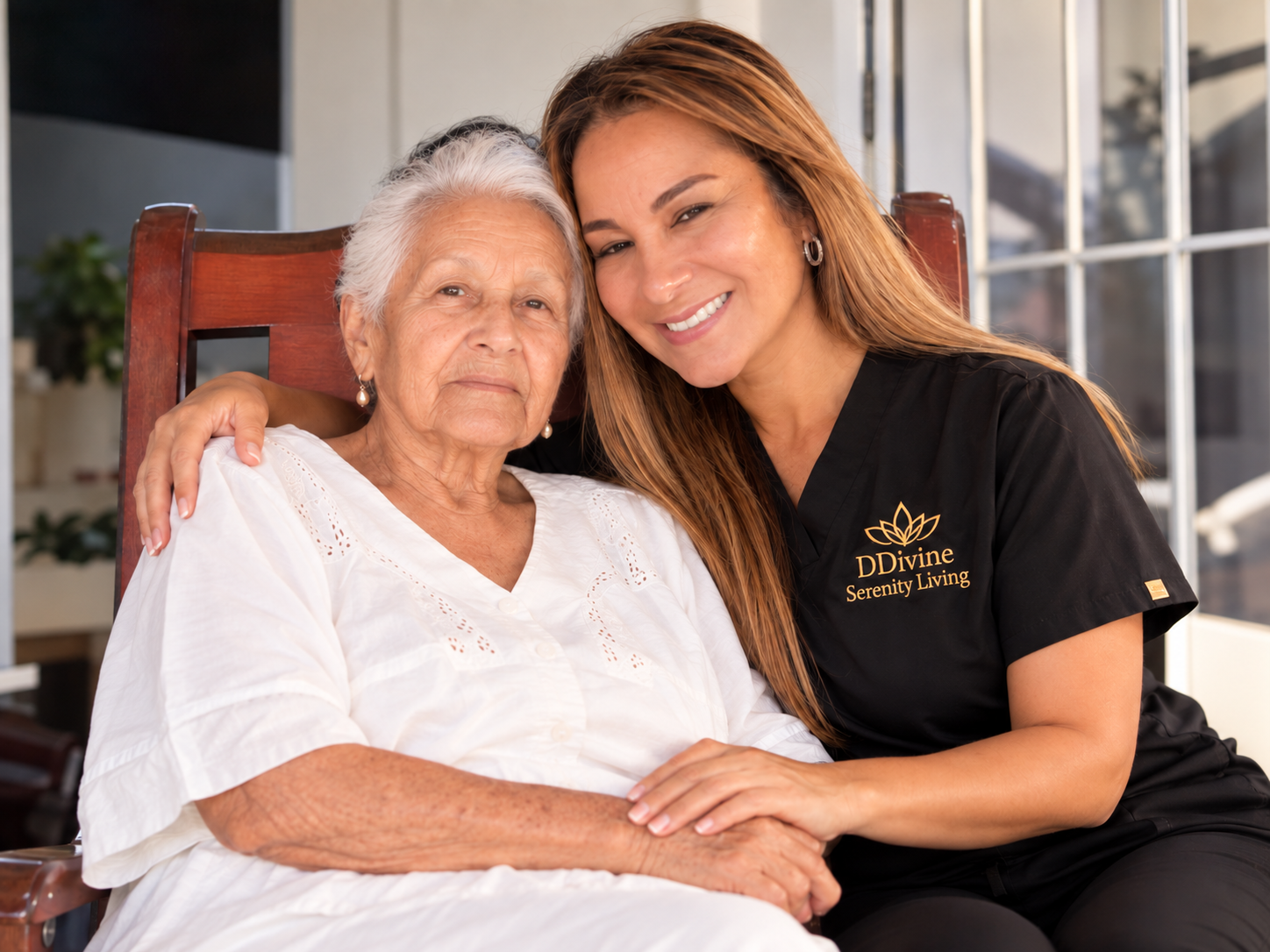 A young woman with long light brown hair and a black uniform with a gold logo saying 'DDivine Serenity Living' is sitting close to an elderly woman with short gray hair, wearing a white blouse, inside a well-lit room with large windows. They are smiling and embracing each other.