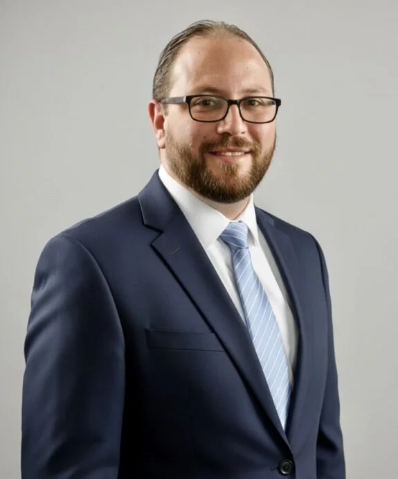 Professional man in a navy suit, light blue tie, white shirt, glasses, smiling, gray background.