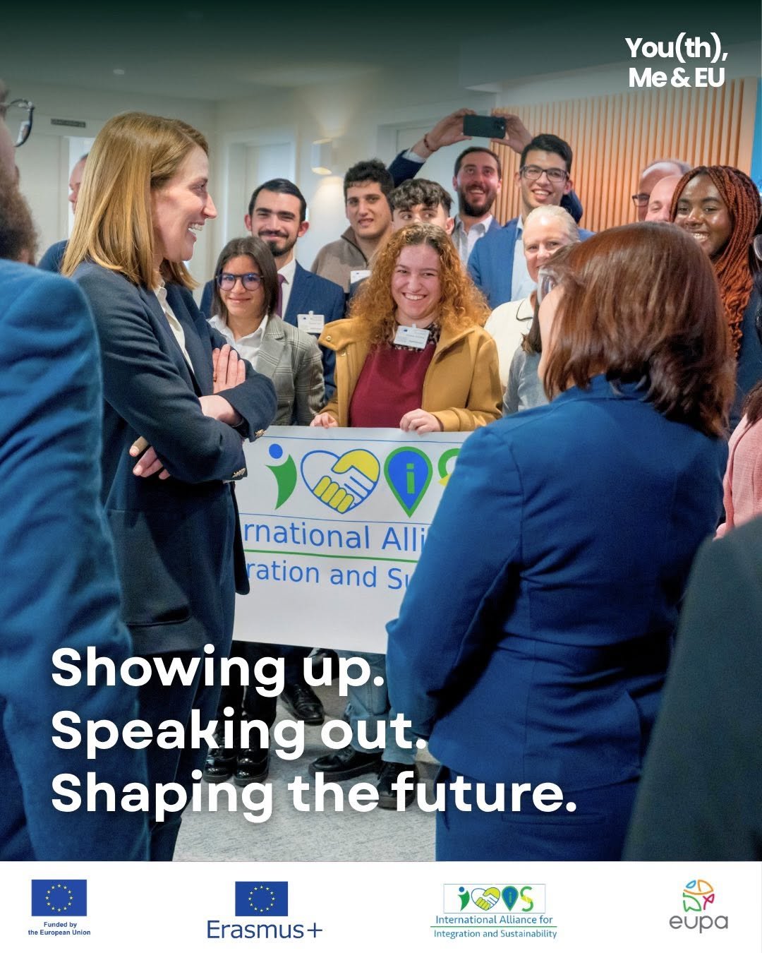 A diverse group of people gathered around two women holding a sign related to the International Alliance for Integration and Sustainability at an event, with some people smiling and one person taking a photo. The background shows a modern indoor setting.