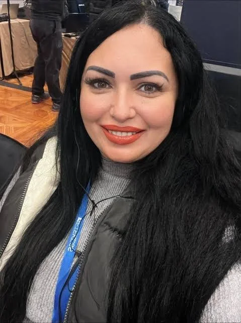A woman with long black hair, smiling with bright makeup and red lipstick, wearing a gray sweater and a black jacket, sitting indoors at an event or conference.