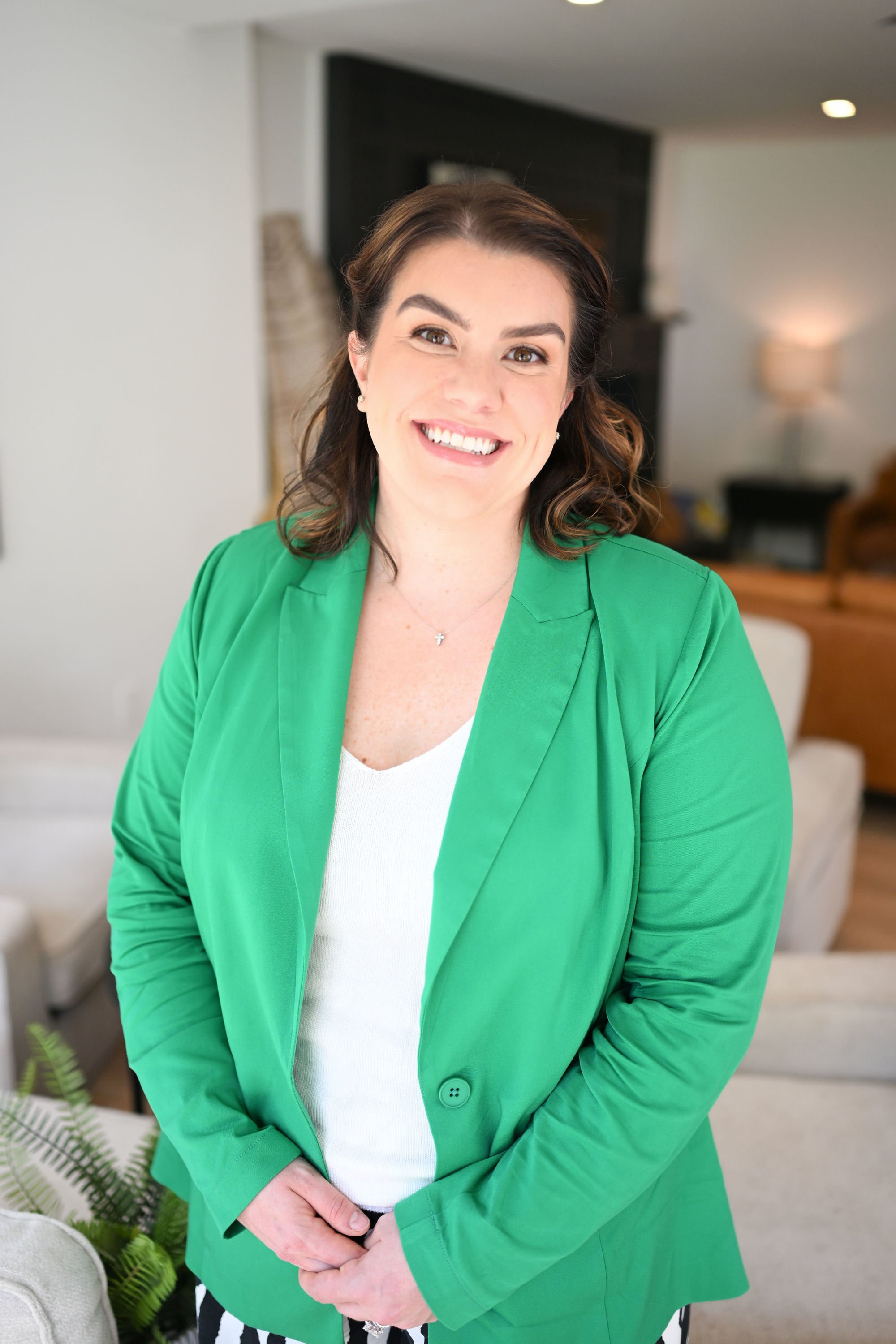 A woman with brown hair smiling, wearing a green blazer and a white top, standing in a living room with a black fireplace, a lamp, and beige furniture in the background.