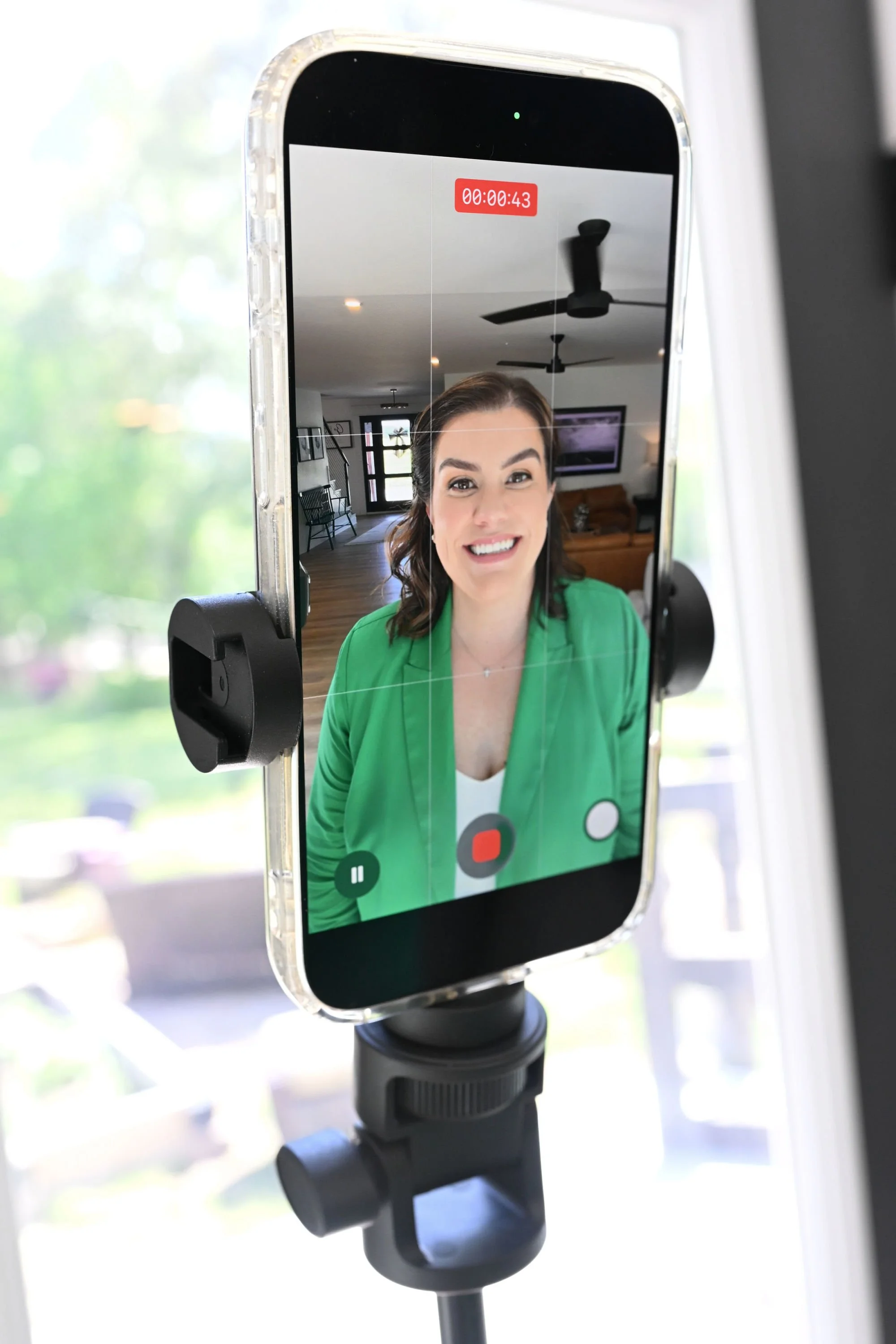 A smartphone mounted on a tripod showing a woman with brown hair, smiling, and wearing a green blazer in a well-lit, modern living room.