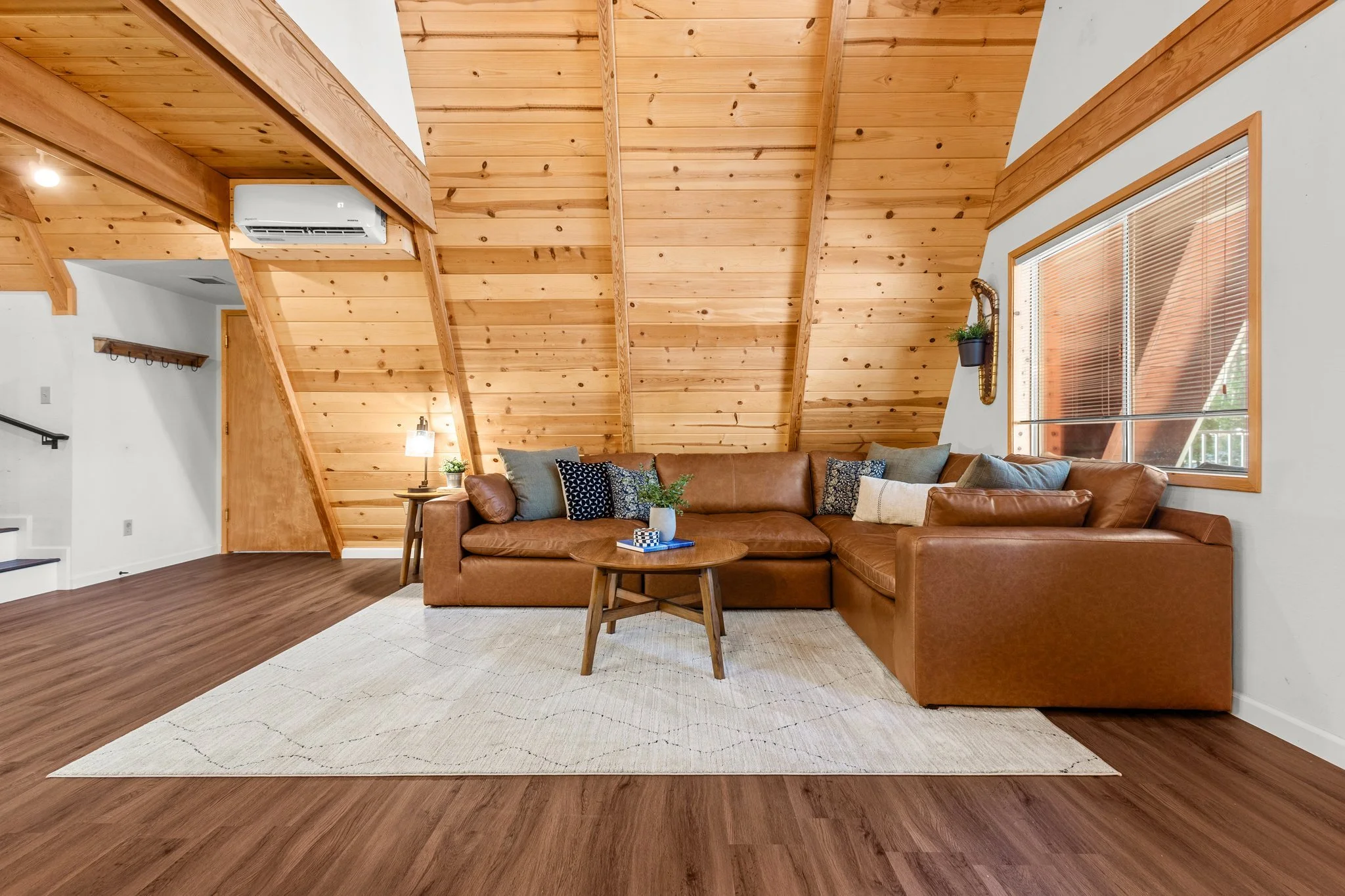 Interior living room with wooden vaulted ceiling, leather sofa with throw pillows, coffee table with decorative items, window with blinds, and a white area rug on wooden floors.