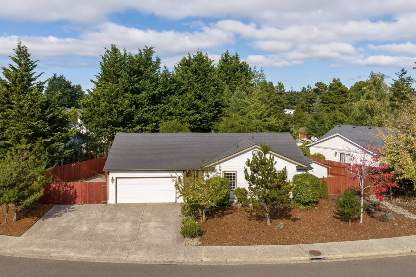 A suburban house with white siding, a gray roof, a two-car garage, surrounded by trees and shrubs, with a red wooden fence on the left, a concrete driveway, and a landscaped yard with mulch and small trees.