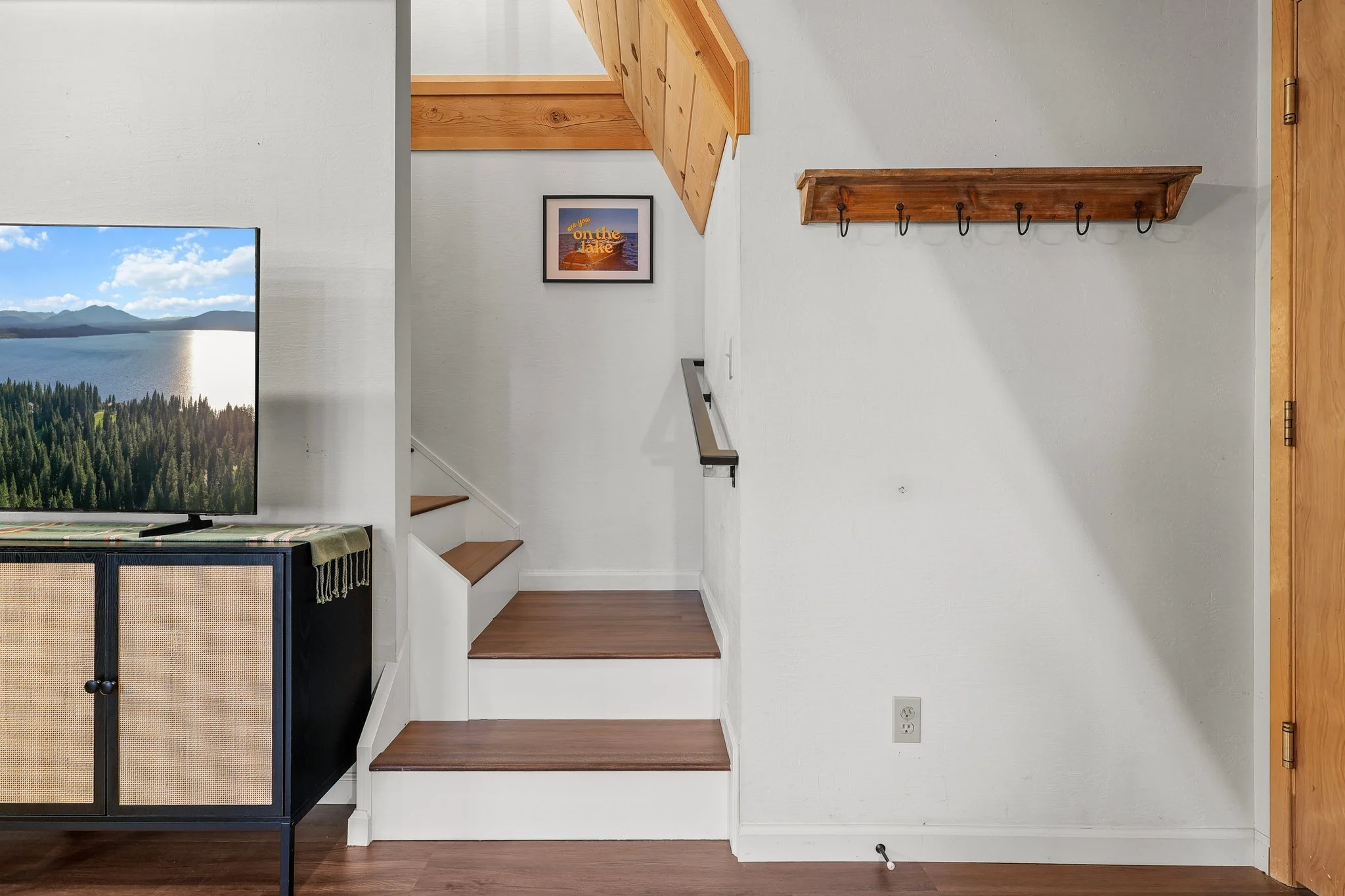 Interior of a home showing part of a living room and a staircase. The room has white walls, wooden stairs, a wooden coat rack with hooks, a framed picture, a TV, and a cabinet. There's an electrical outlet on the wall near the floor.