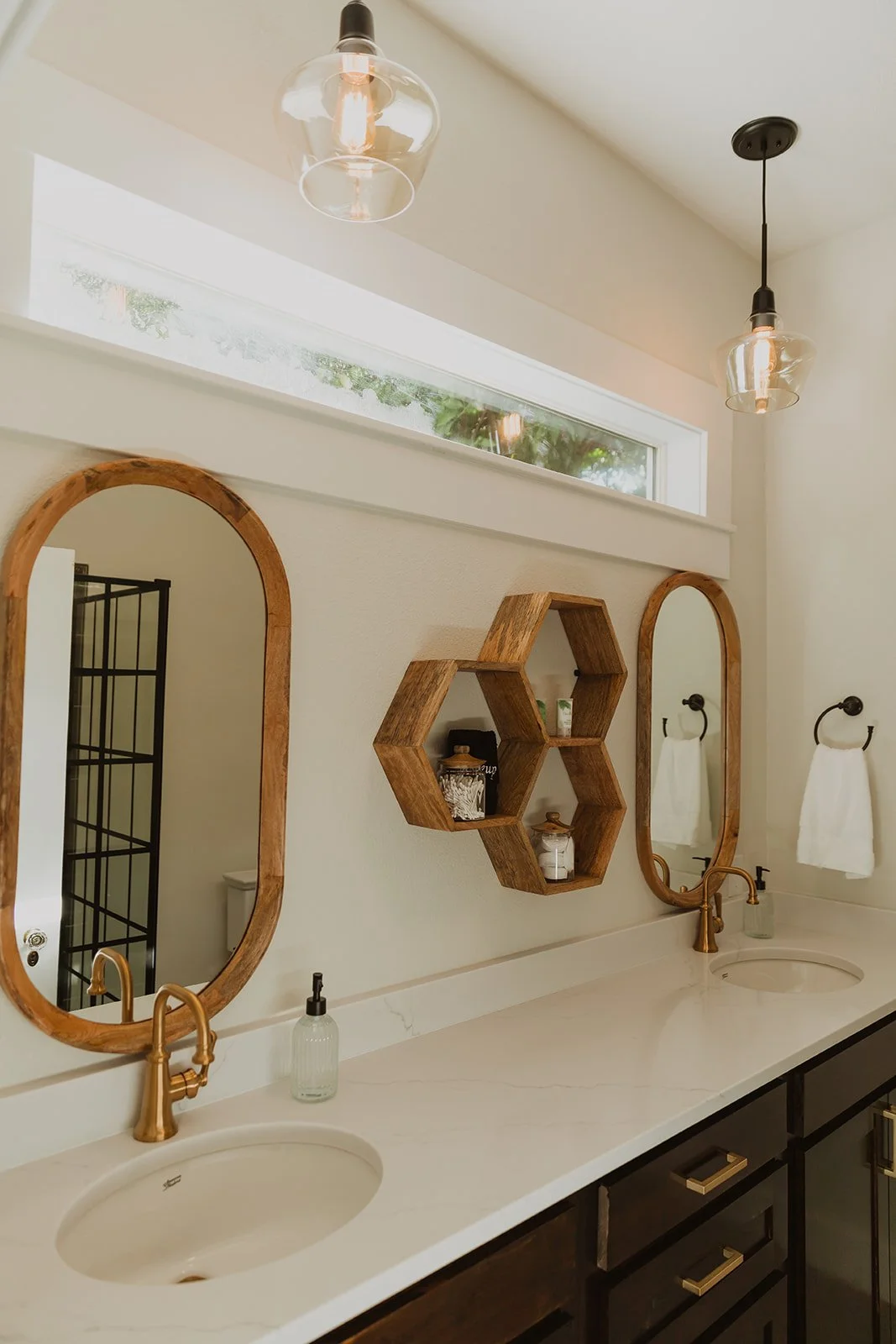 Modern bathroom with double sinks, round mirrors with wooden frames, hexagonal wooden shelves, white countertops, black cabinets, brass faucets, and black light fixtures.
