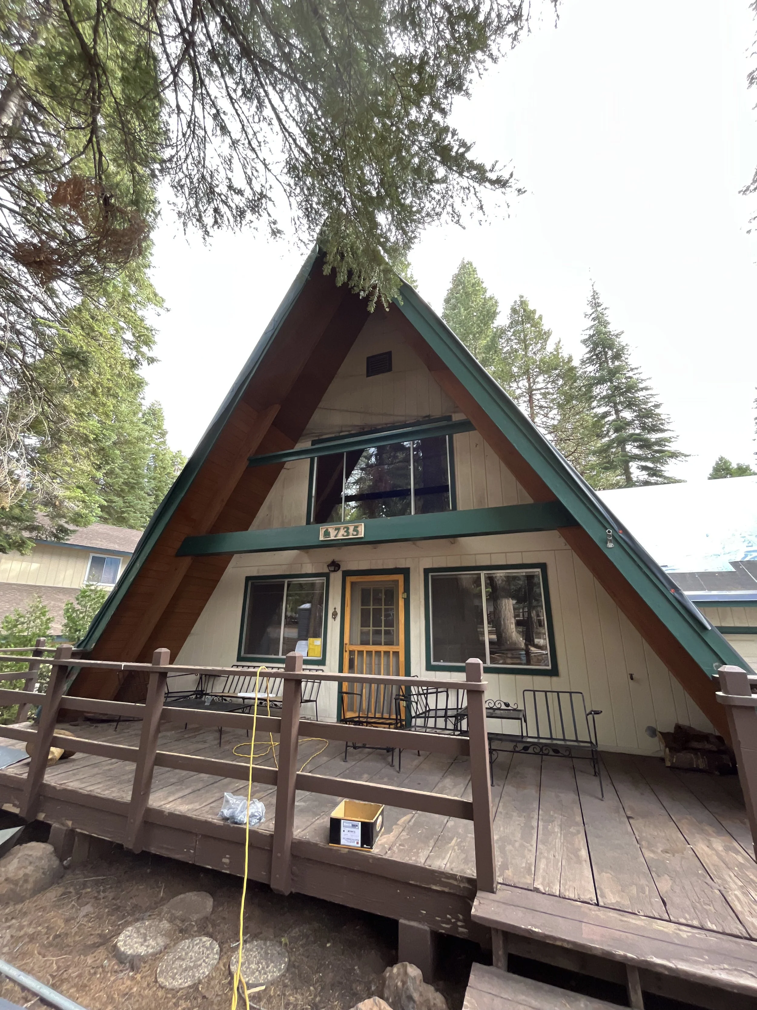 The photo shows an A-frame house with a wooden deck. There are some chairs and tables on the deck, and construction tools and materials are present, indicating ongoing work. Tall trees surround the house, and the sky above is overcast.